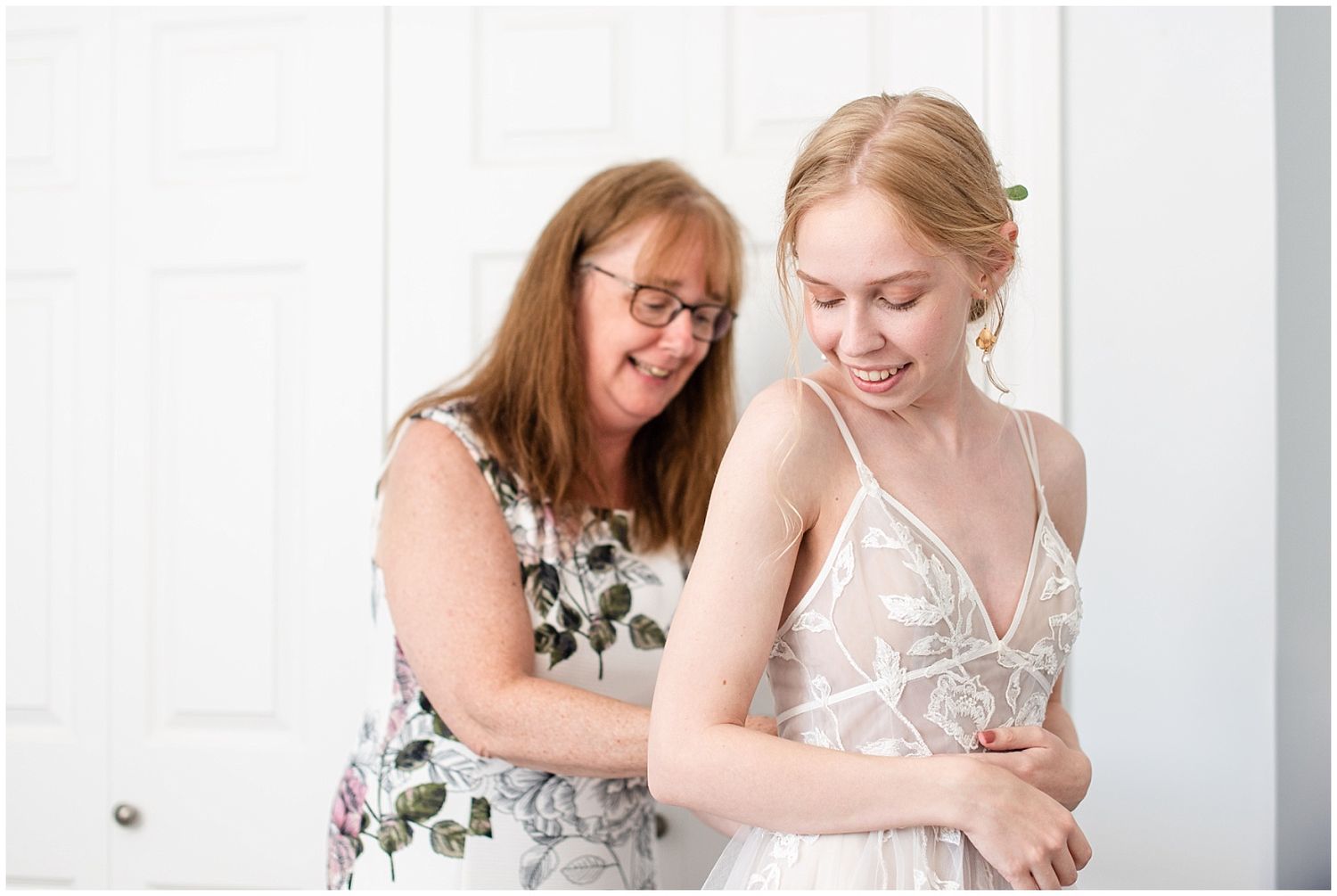 Mother helping bride into lace wedding dress in bright white room during bridal preparations.