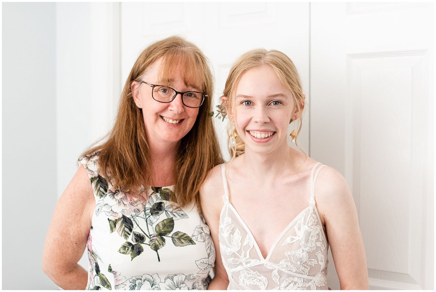 Two smiling women pose together, one in floral dress with glasses, the other in a white lace bridal gown with floral hair accessories.