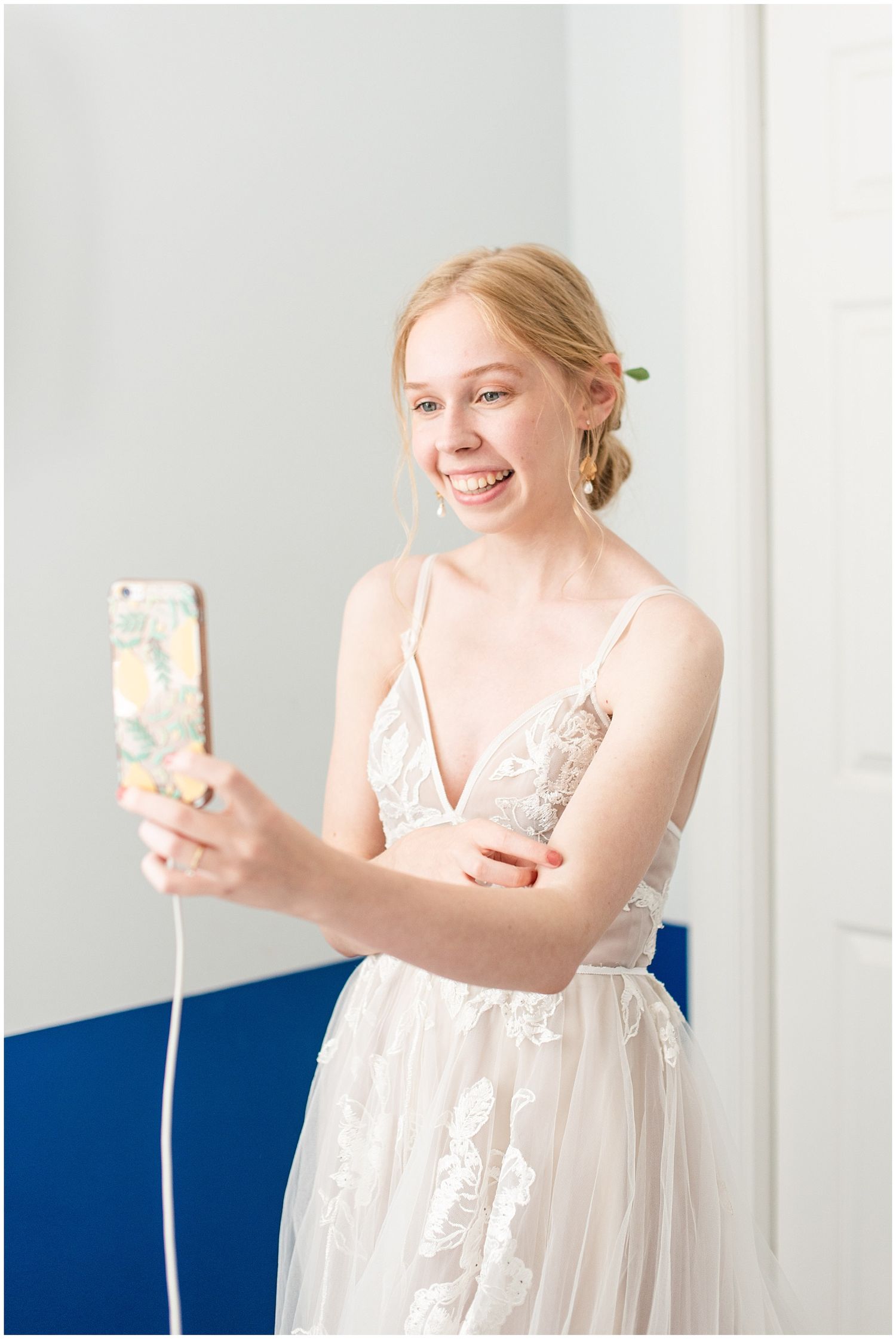 Smiling bride in lace wedding dress taking a selfie with floral phone case in bright white room.