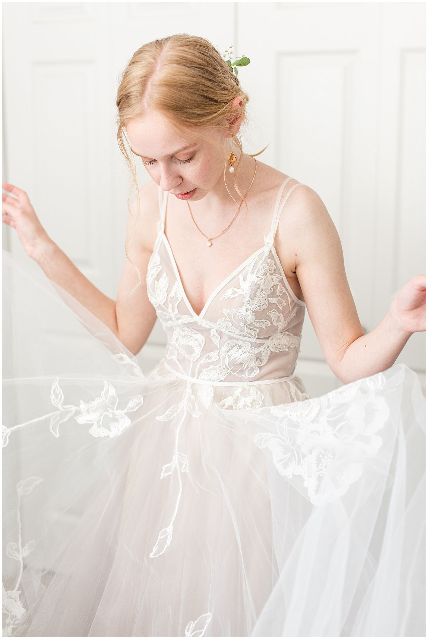 Bride in lace tulle wedding gown holding dress skirt, wearing pearl earrings and delicate necklace with floral hair piece.