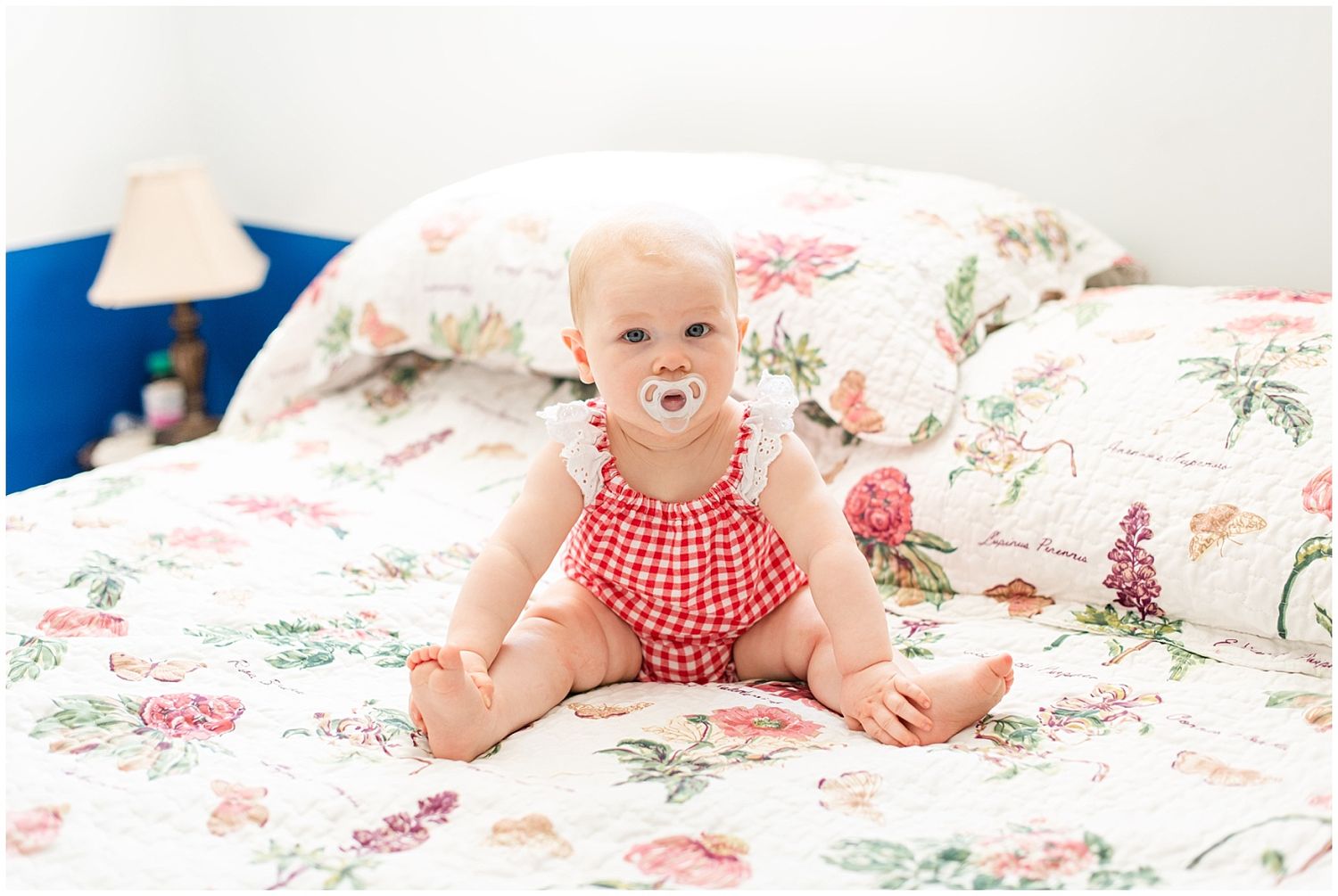 Smiling baby in red gingham romper sitting on floral bedding with a pacifier.