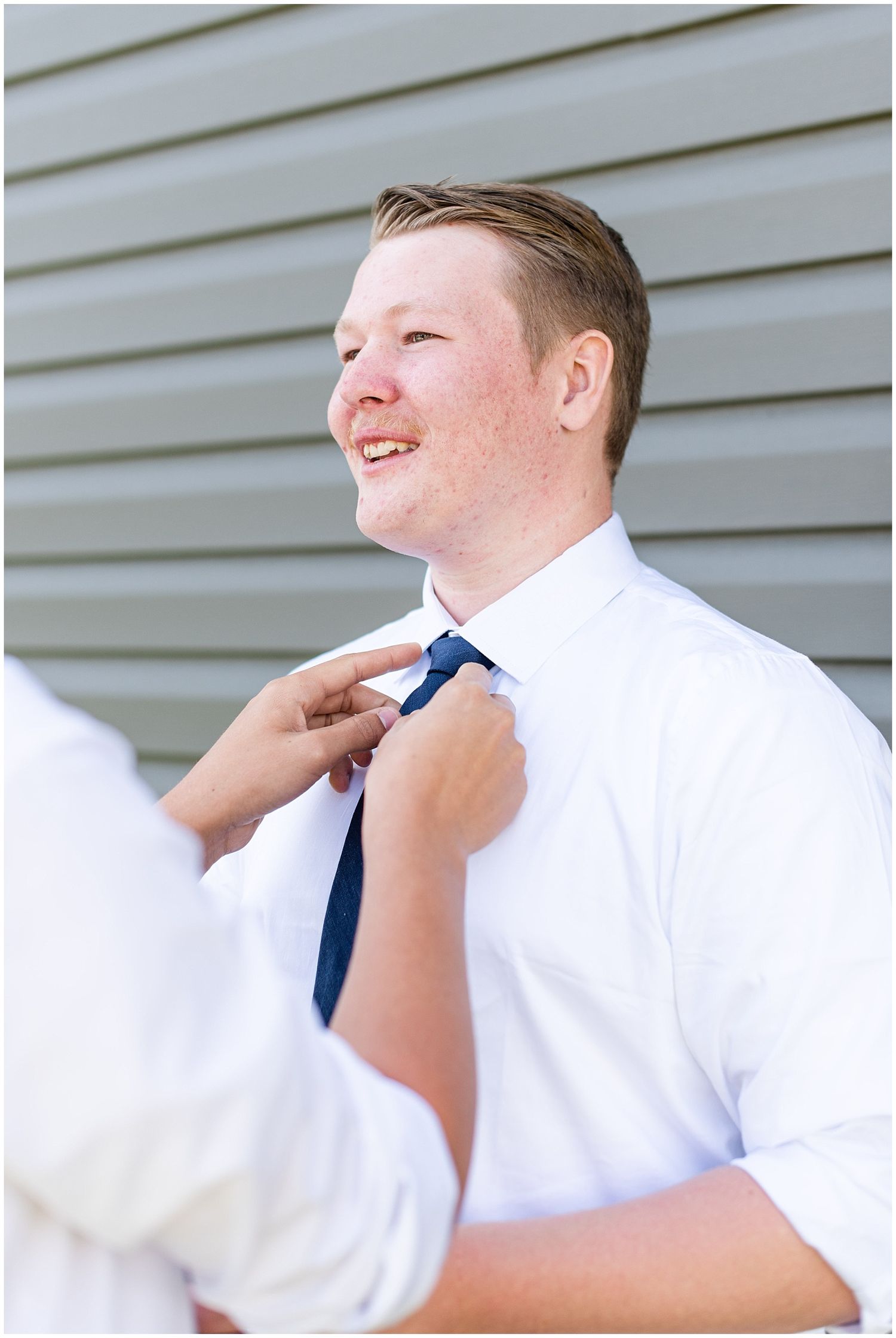 Groom smiling as someone adjusts his navy blue tie, wearing a white dress shirt outdoors.