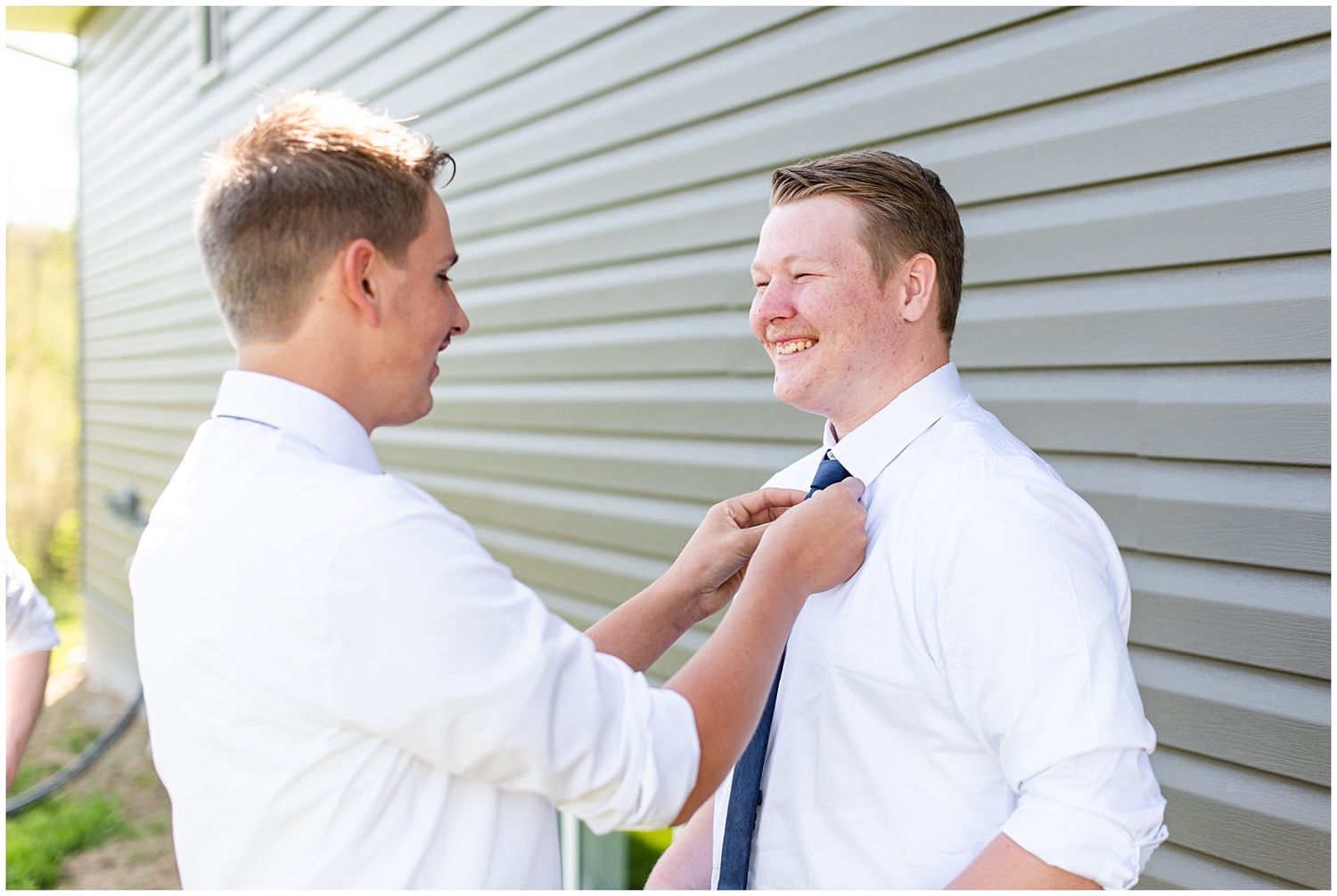 Groomsman helping groom adjust his blue tie outside before a wedding ceremony.