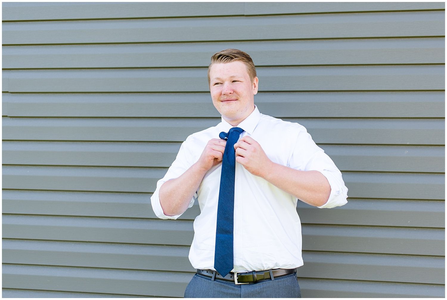 Man in white dress shirt adjusting navy blue tie outdoors against gray siding.