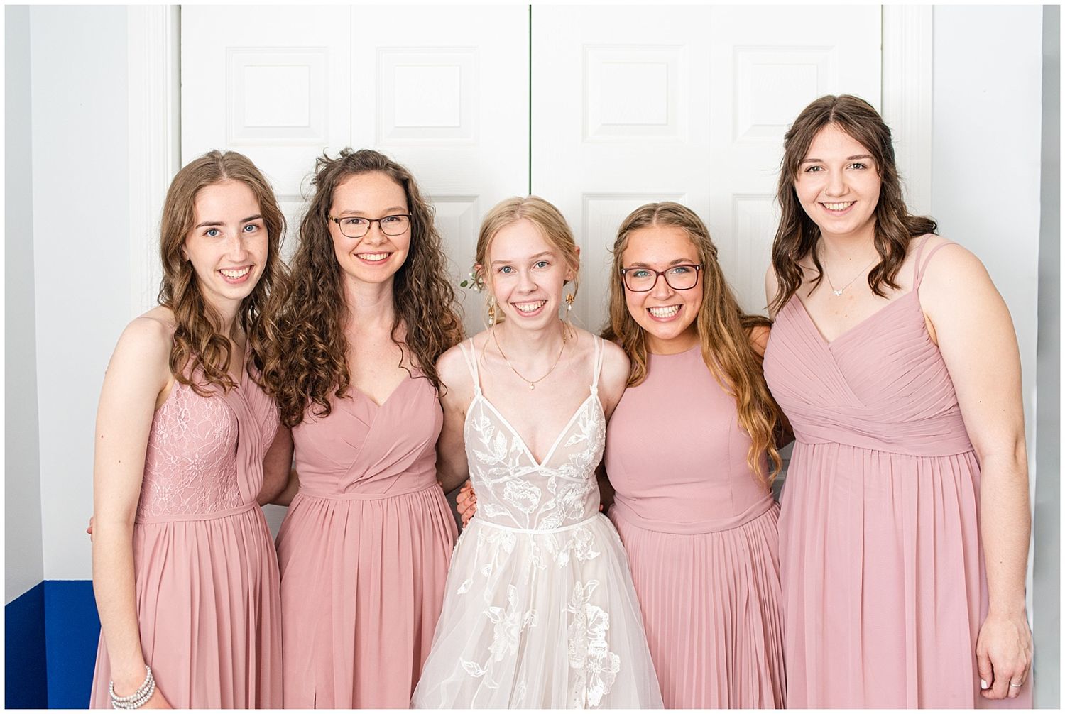 Bride in white lace gown poses with four bridesmaids in dusty rose dresses indoors.