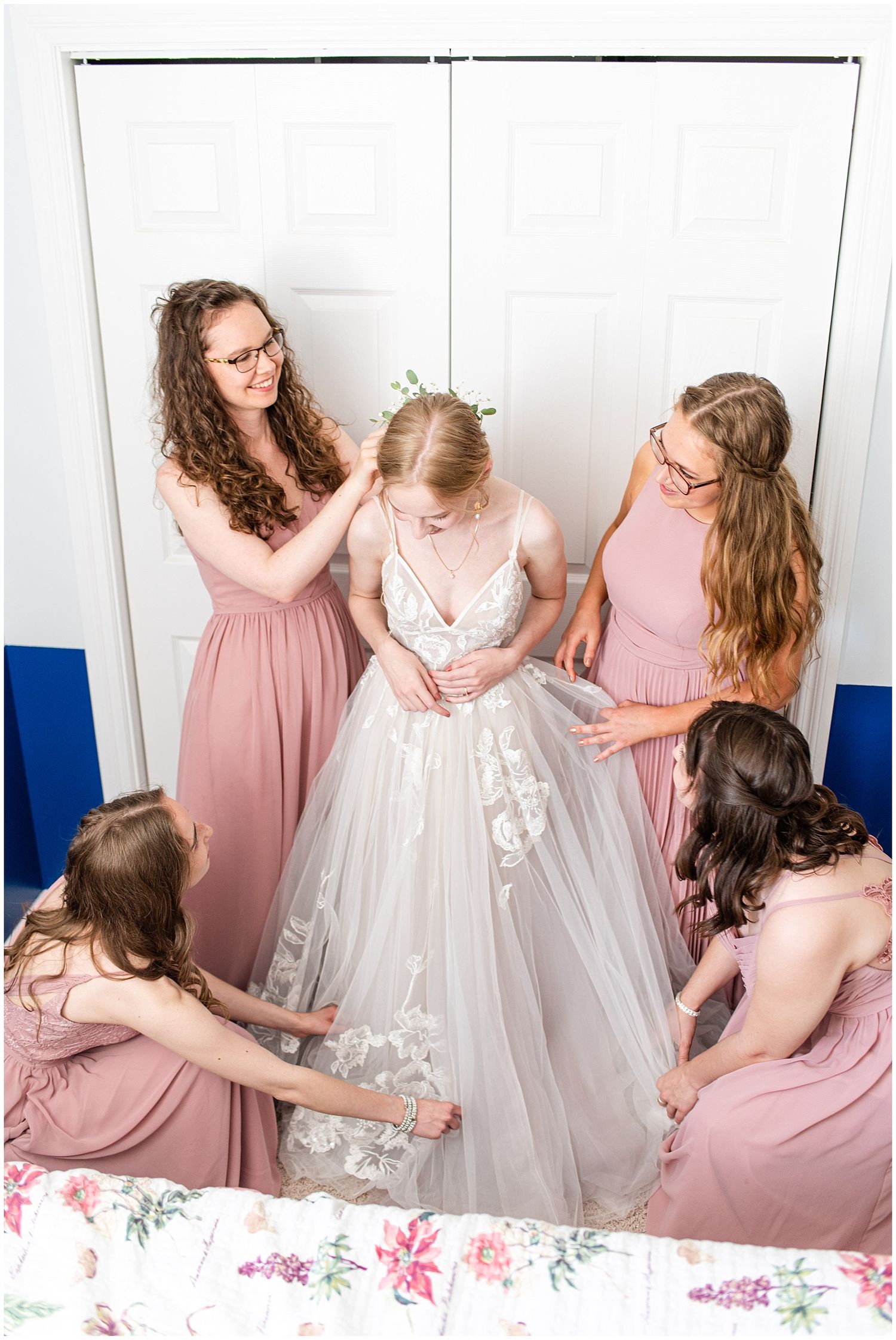 Bridesmaids in dusty rose gowns help bride into white floral wedding dress before ceremony.