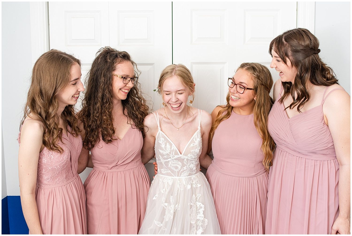 Bride in white lace gown laughing joyfully with four bridesmaids in dusty rose dresses indoors.