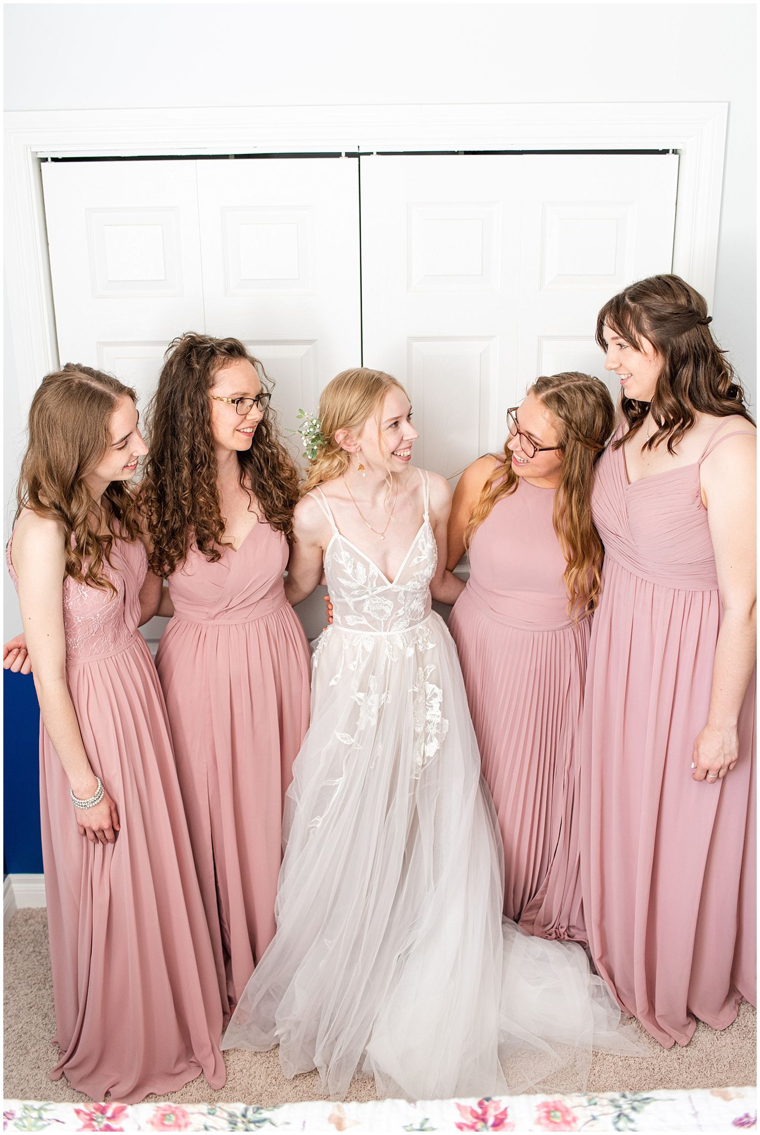 Bride in a flowy white gown surrounded by four bridesmaids in mauve pink dresses smiling together.