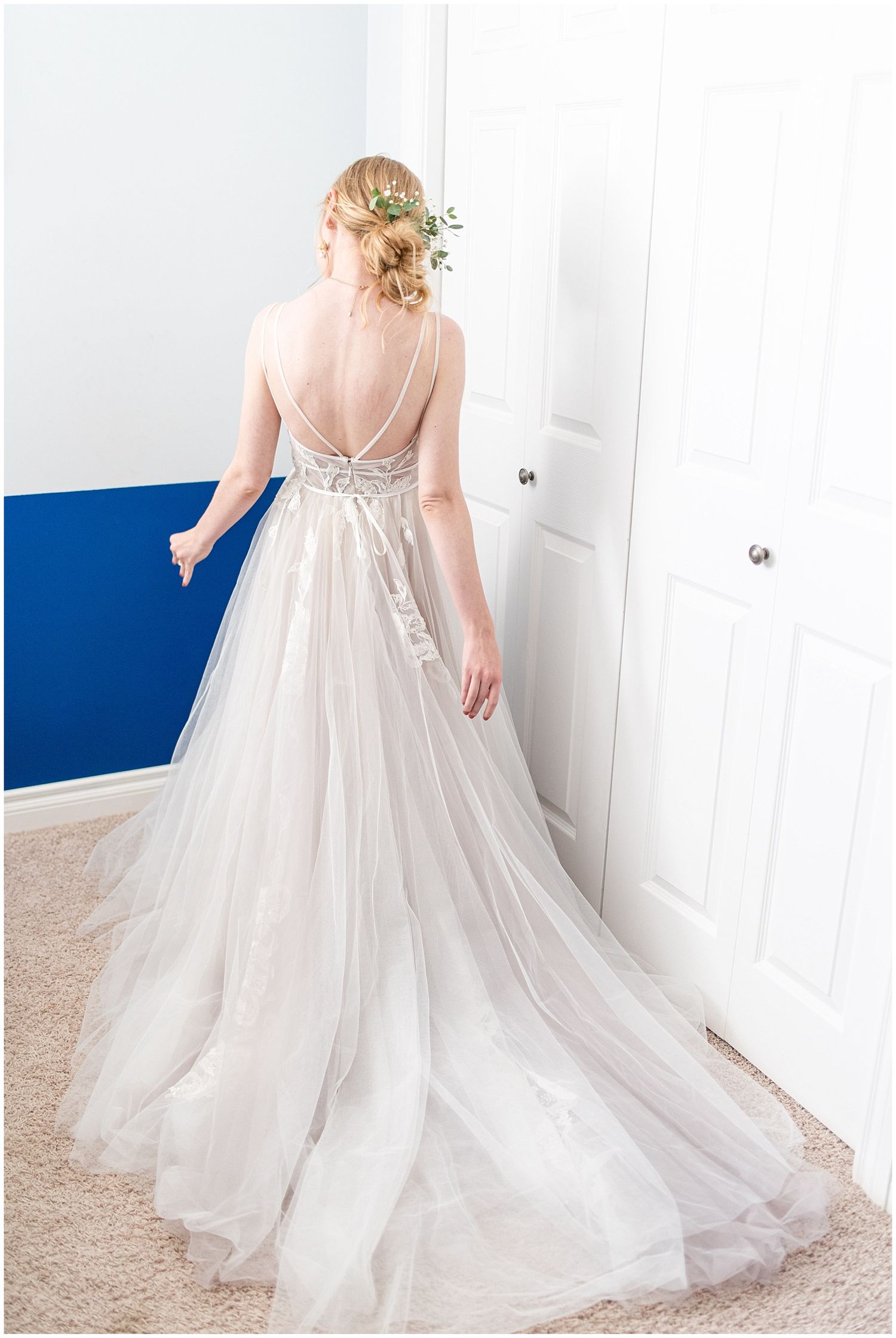 Bride in a backless tulle wedding gown with floral hair accessory, standing near a white door with blue wall.