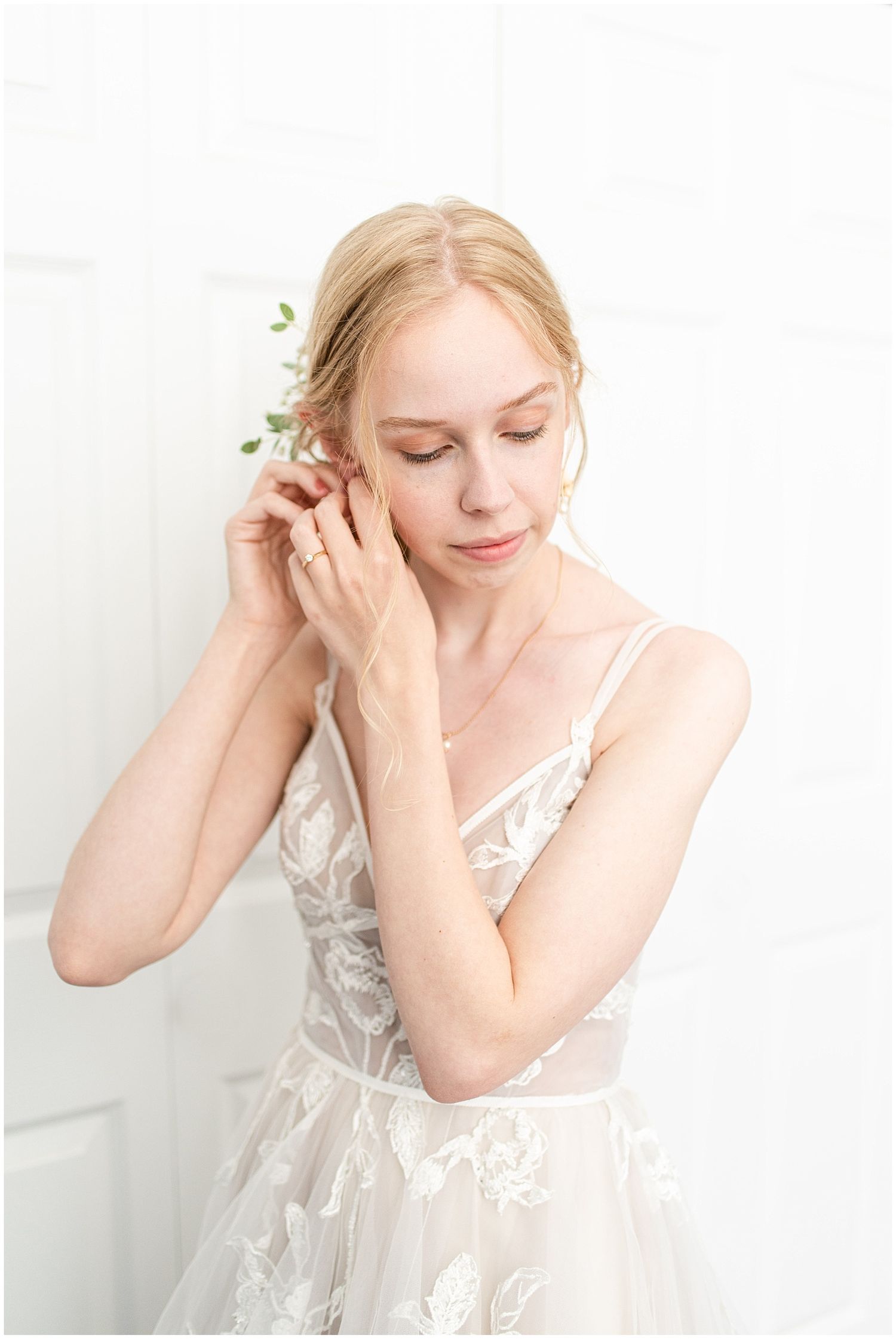 Blonde bride in floral lace wedding dress holding a small green sprig near her ear.