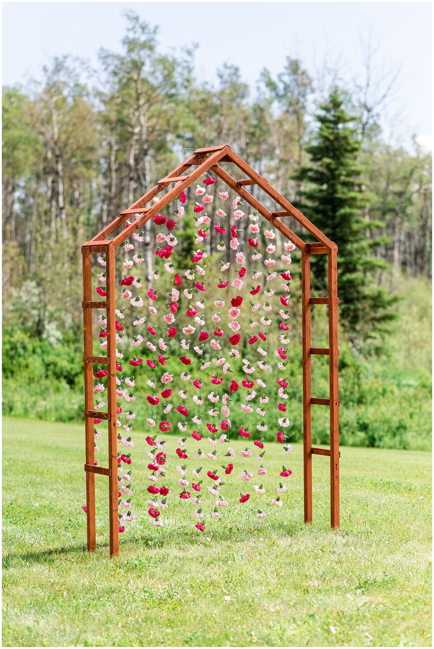 A rustic wooden arch adorned with cascading pink and red floral garlands stands in a lush green outdoor setting.