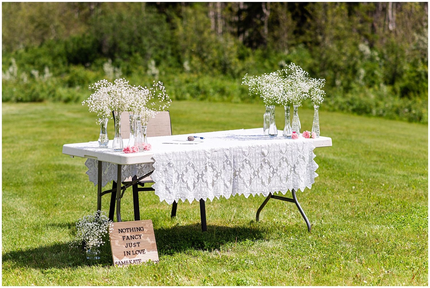Outdoor wedding table with white lace cloth, baby's breath in glass bottles, on a sunny green lawn.