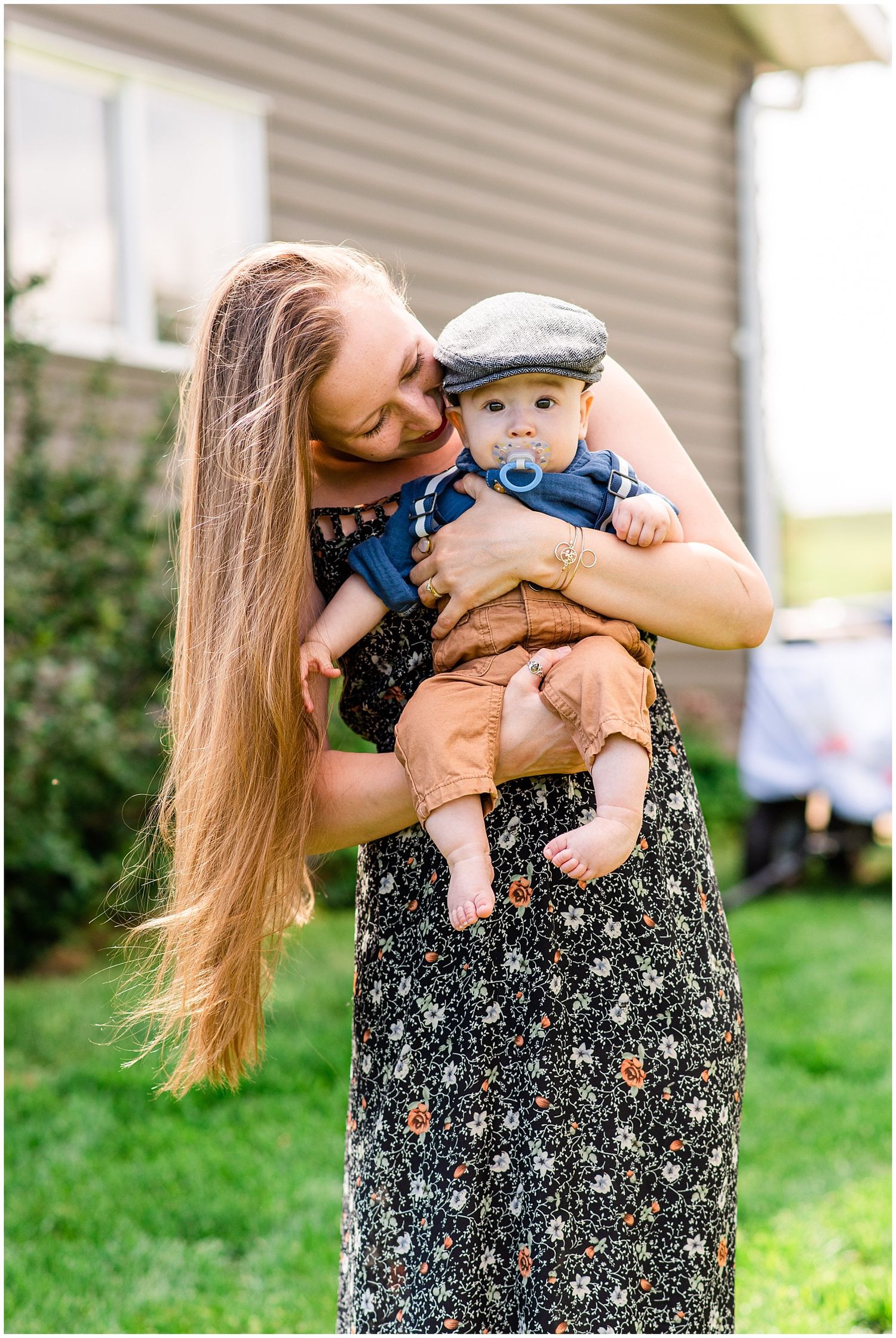 Mother with long blonde hair holding her baby boy in a newsboy cap and blue outfit outdoors.