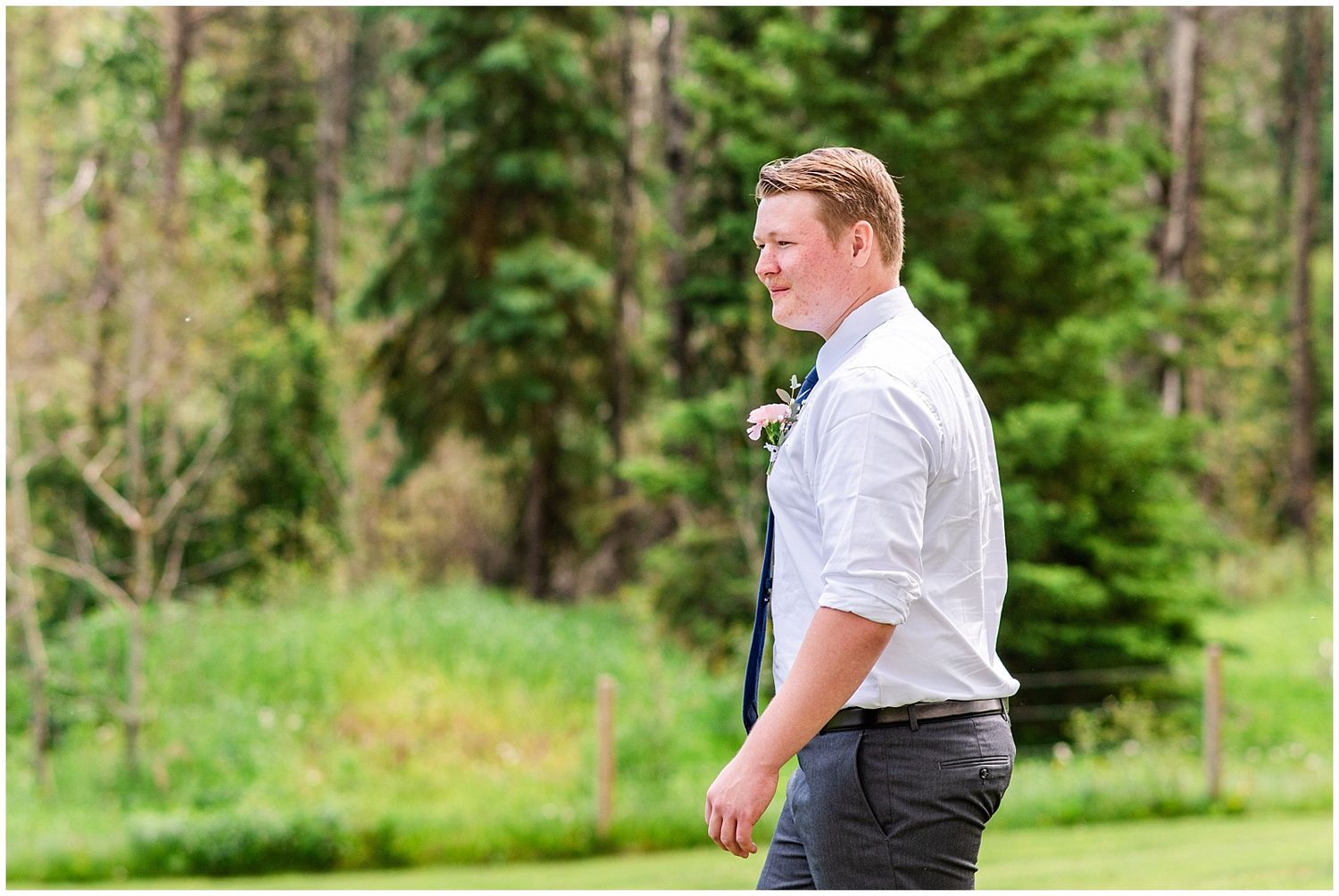 Groom in white dress shirt and tie standing outdoors in front of lush green trees.
