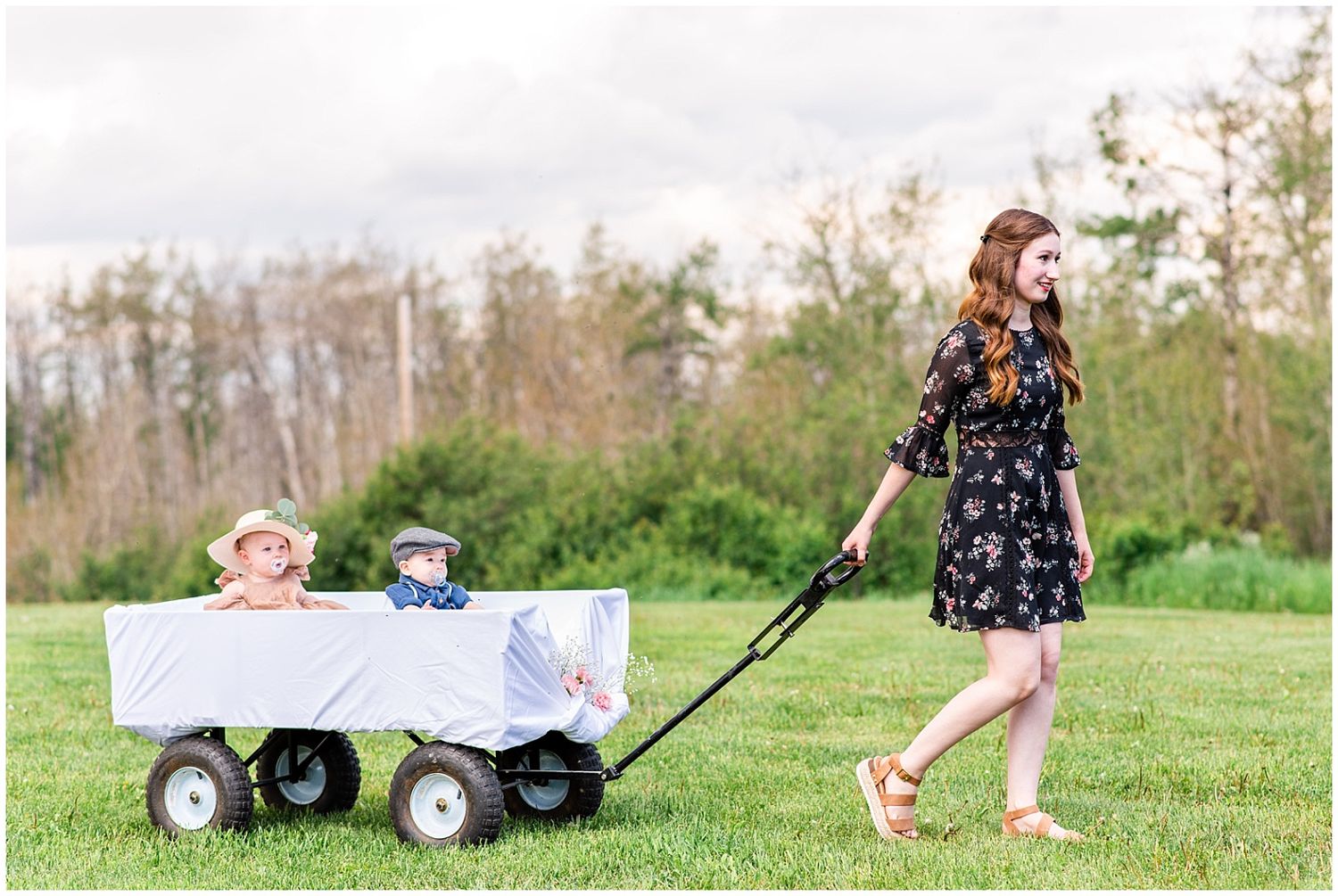 Woman in floral dress pulling two young children in a white wagon across a grassy outdoor wedding venue.