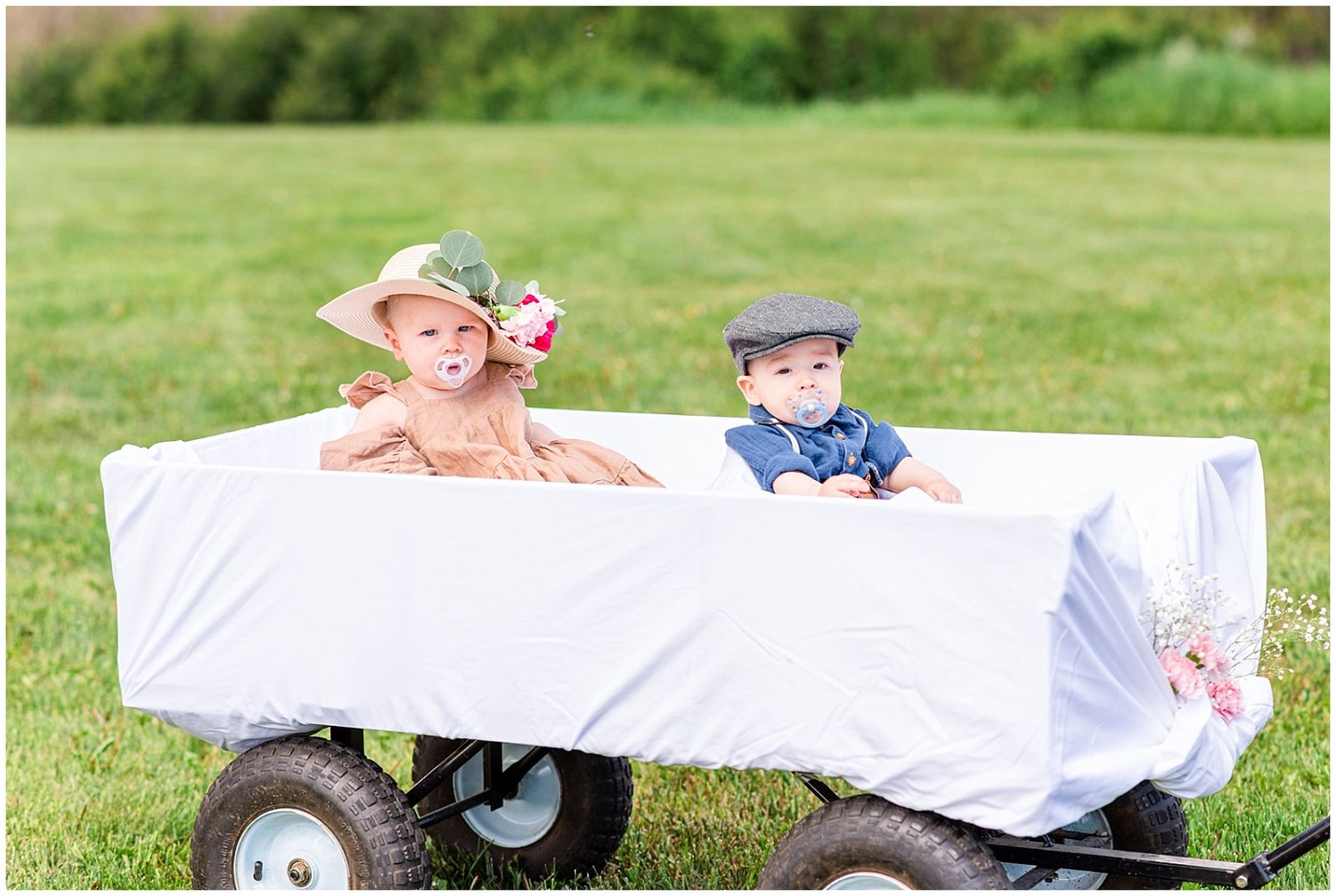Two young children in formal attire ride in a white-draped wagon on green grass.