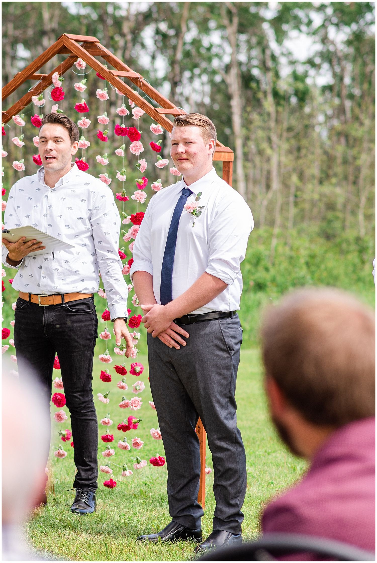 Groom smiling at outdoor wedding ceremony beneath floral arch with red and pink hanging flowers.