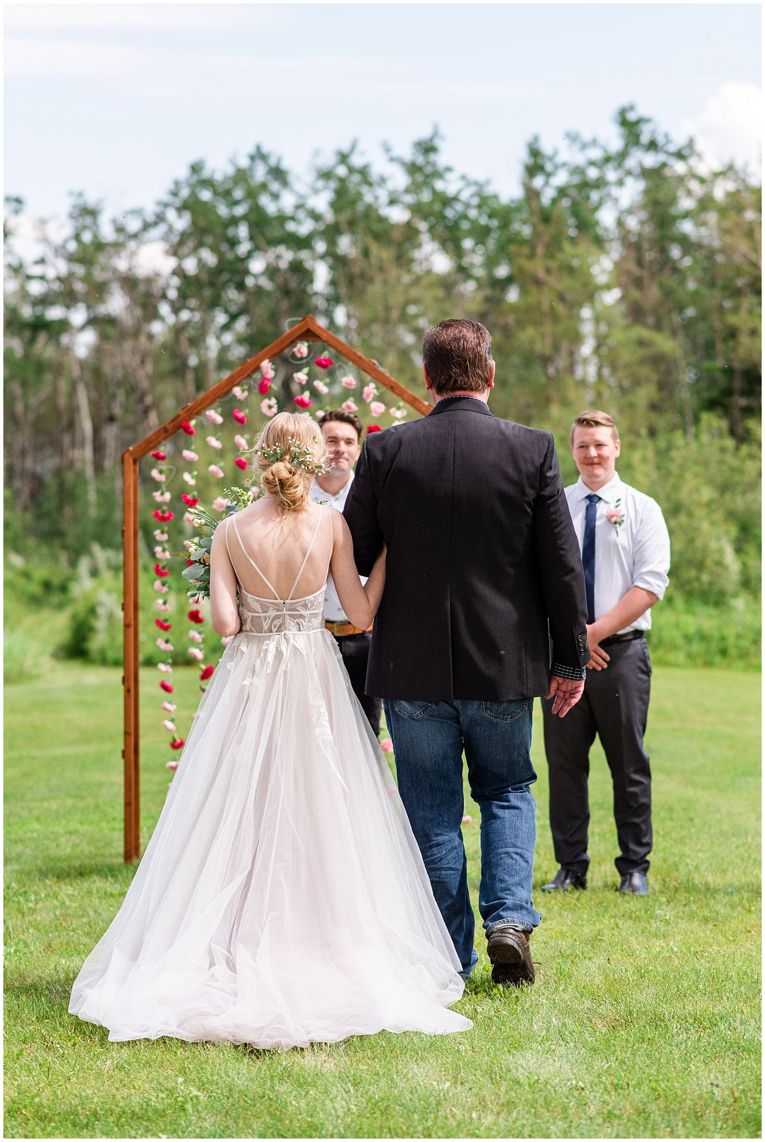 Bride in backless gown walks toward floral arch with groom during outdoor wedding ceremony.