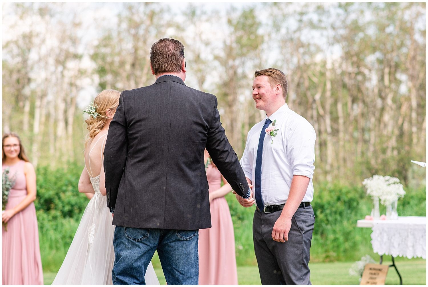 Groom smiling as bride is walked down the aisle at an outdoor wedding ceremony with pink bridesmaid dresses.