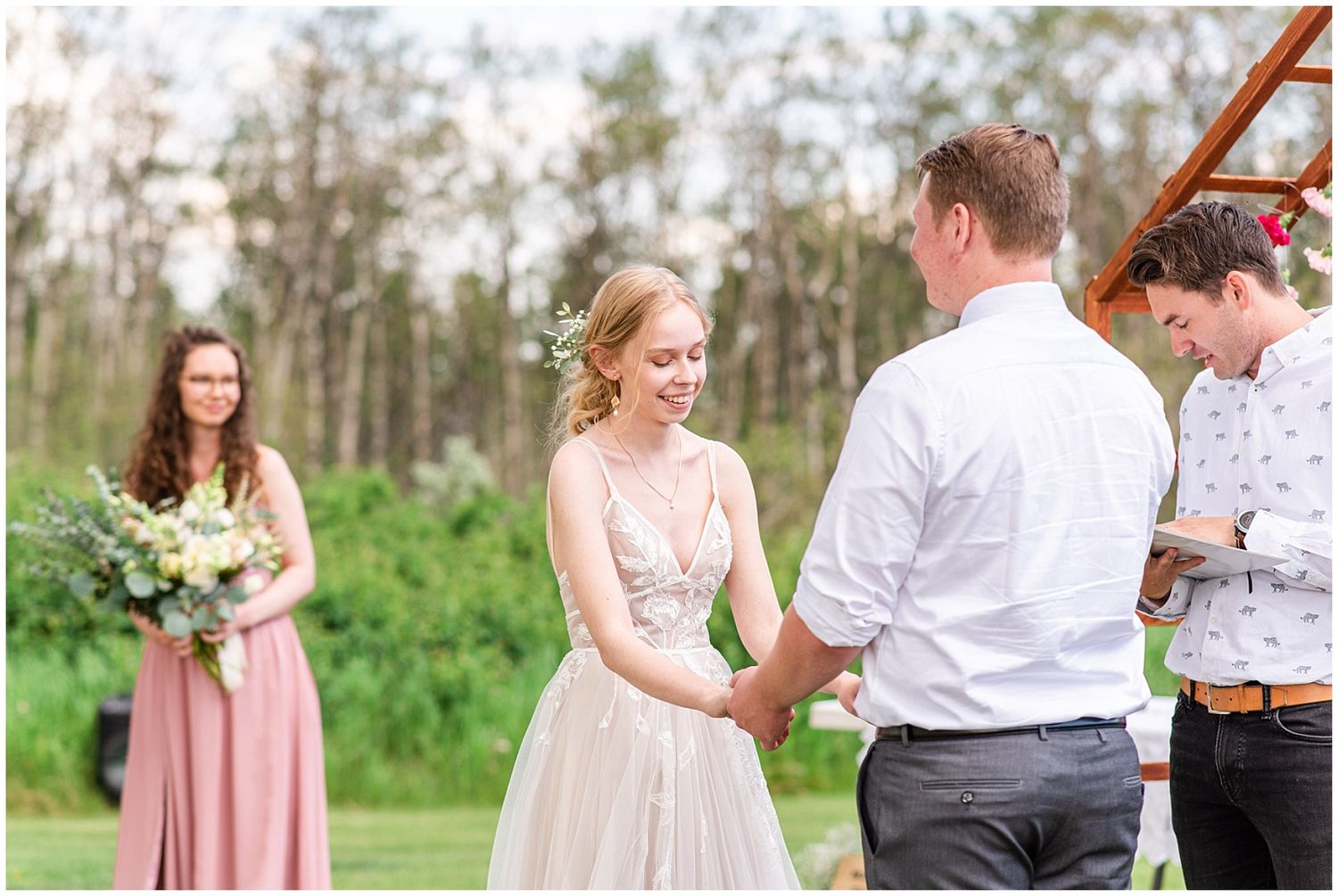Bride and groom holding hands during outdoor wedding ceremony with bridesmaid in pink dress nearby.