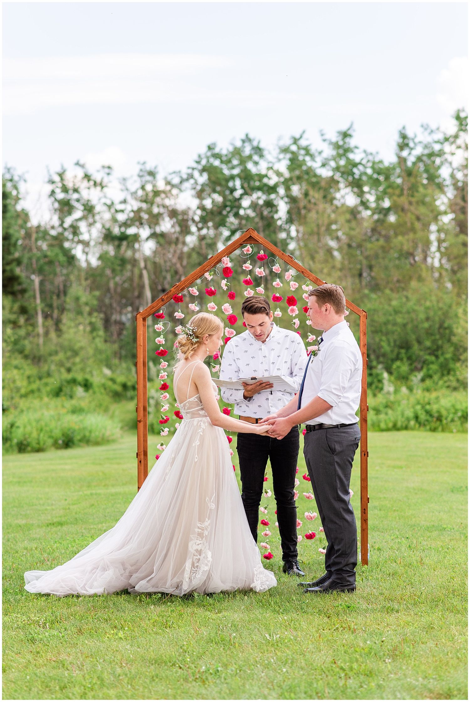 Couple exchanging vows at outdoor wedding altar decorated with red flowers and wooden arch structure.