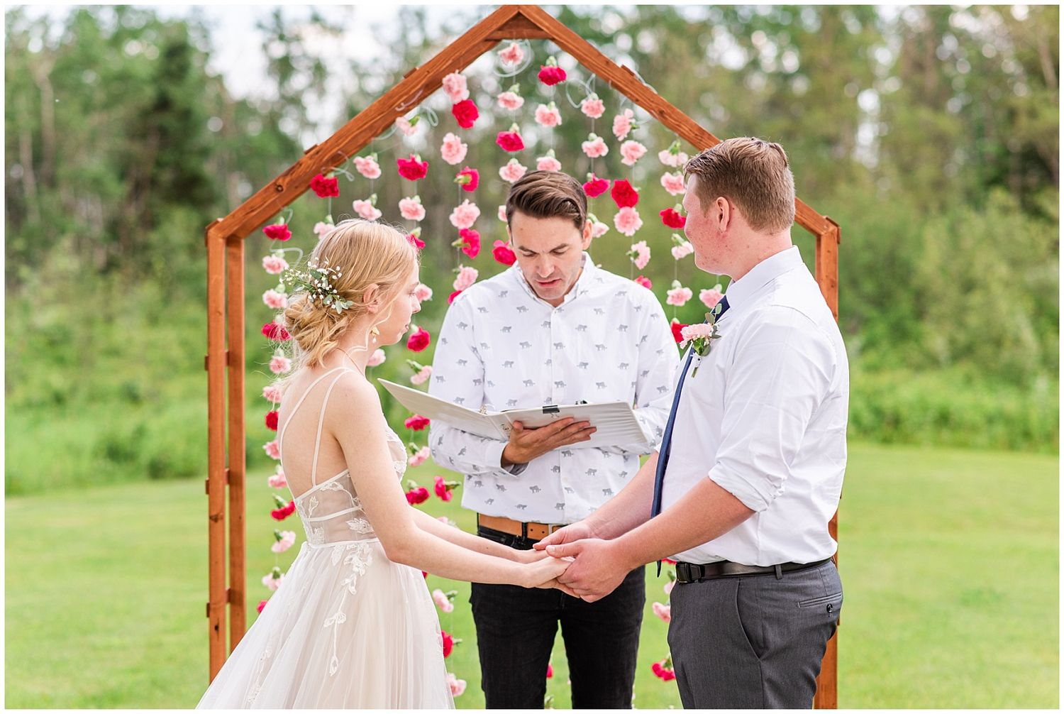 Bride and groom exchange vows under wooden arch adorned with pink and red flowers.