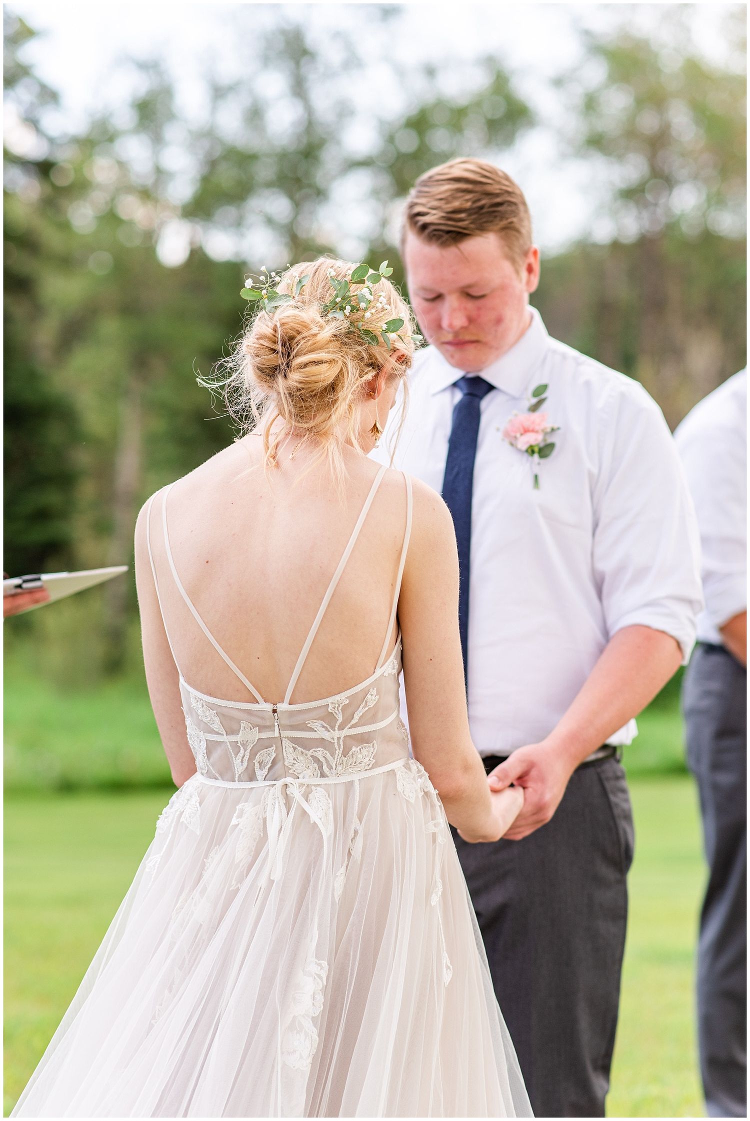 Bride in backless lace wedding gown holds grooms hands during outdoor wedding ceremony.