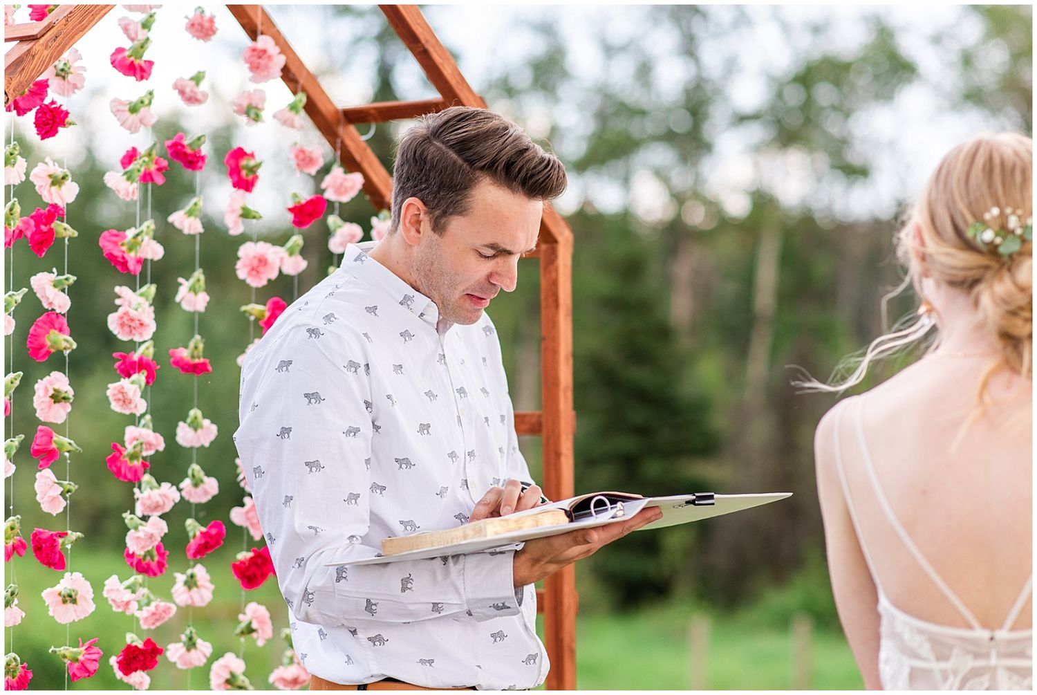 Officiant reads vows from clipboard at flower-adorned wooden arch during outdoor wedding.