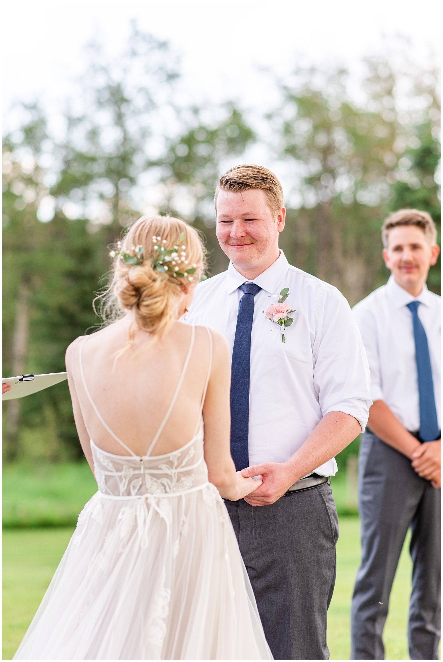 Groom smiling at bride during outdoor wedding ceremony, wearing white shirt and navy tie with floral boutonniere.