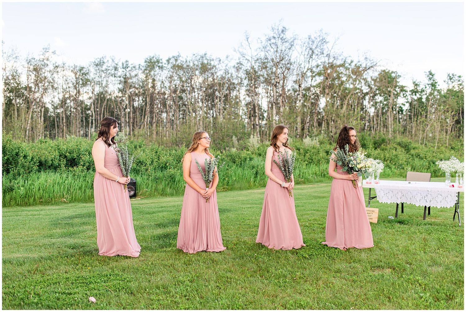 Four bridesmaids in blush pink gowns stand on a green lawn during an outdoor wedding ceremony.