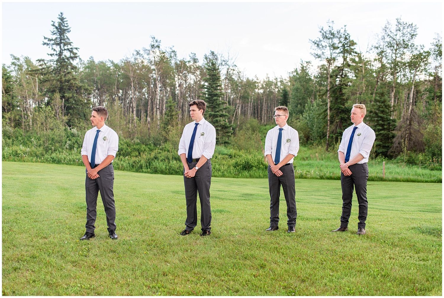 Four groomsmen in white shirts and navy ties stand in a row on a green lawn with trees in the background.