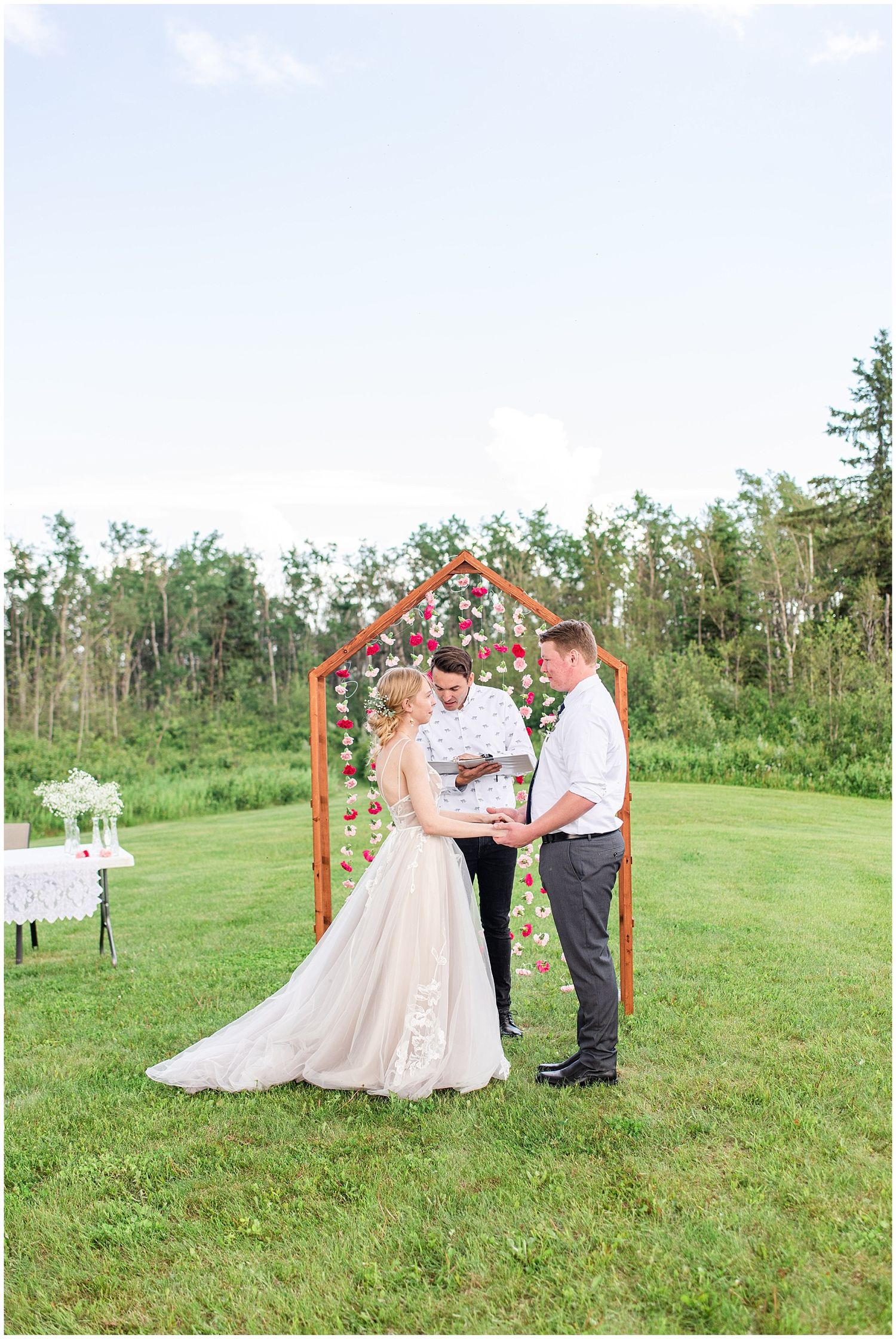 Bride and groom exchange vows outdoors under a wooden arch adorned with red flowers on a lush green lawn.