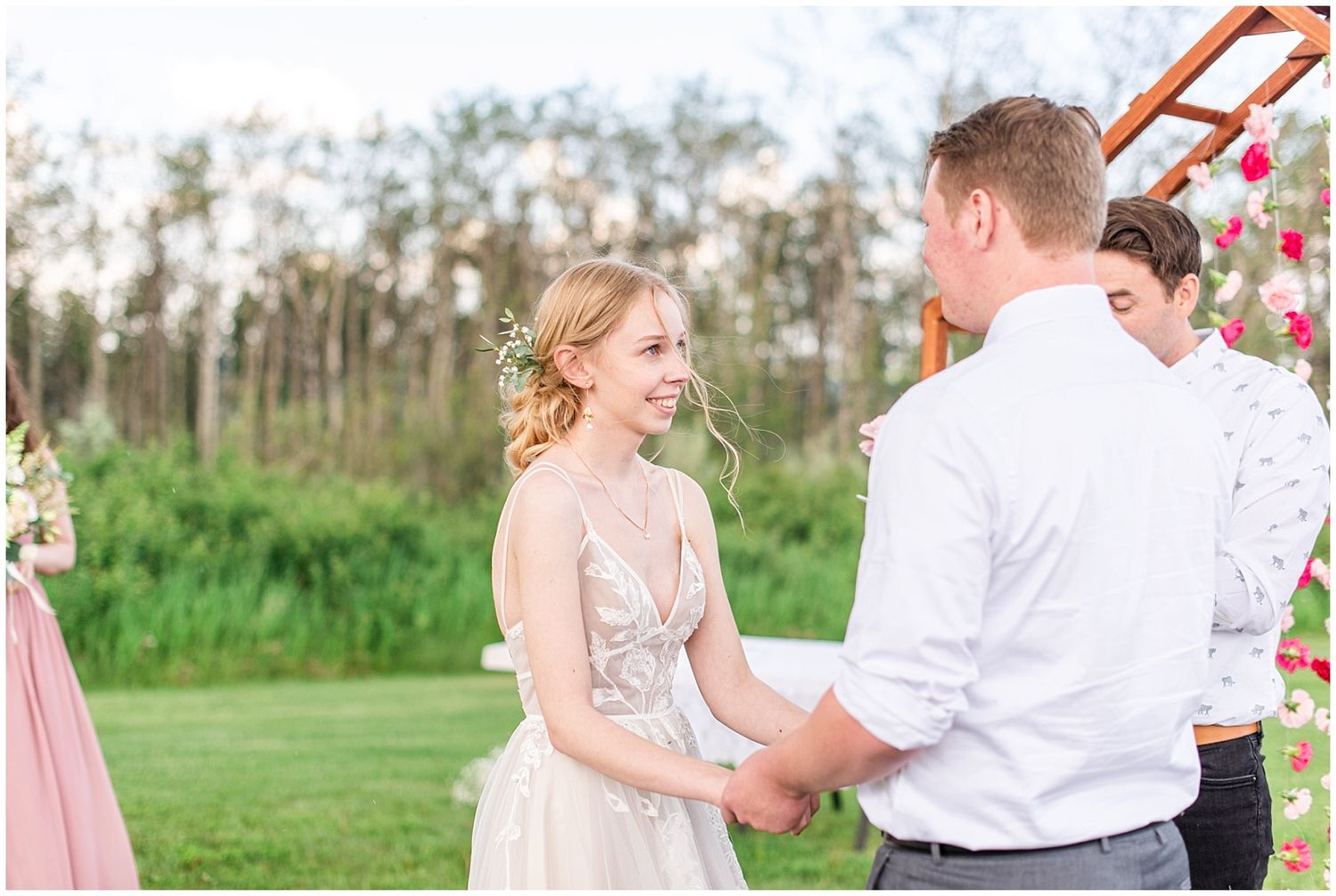 Smiling bride in floral gown holds grooms hands at outdoor wedding ceremony altar.