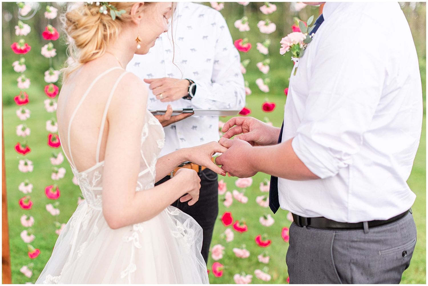 Bride and groom exchange rings at outdoor wedding ceremony with floral backdrop of red and white flowers.