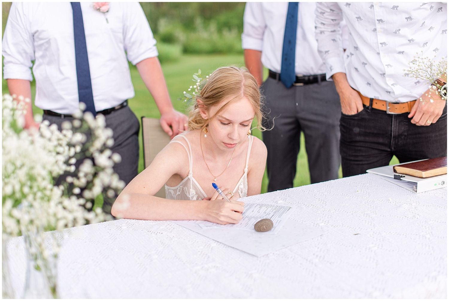 Blonde bride signs wedding documents at a white table outdoors, with groomsmen in navy ties standing nearby.