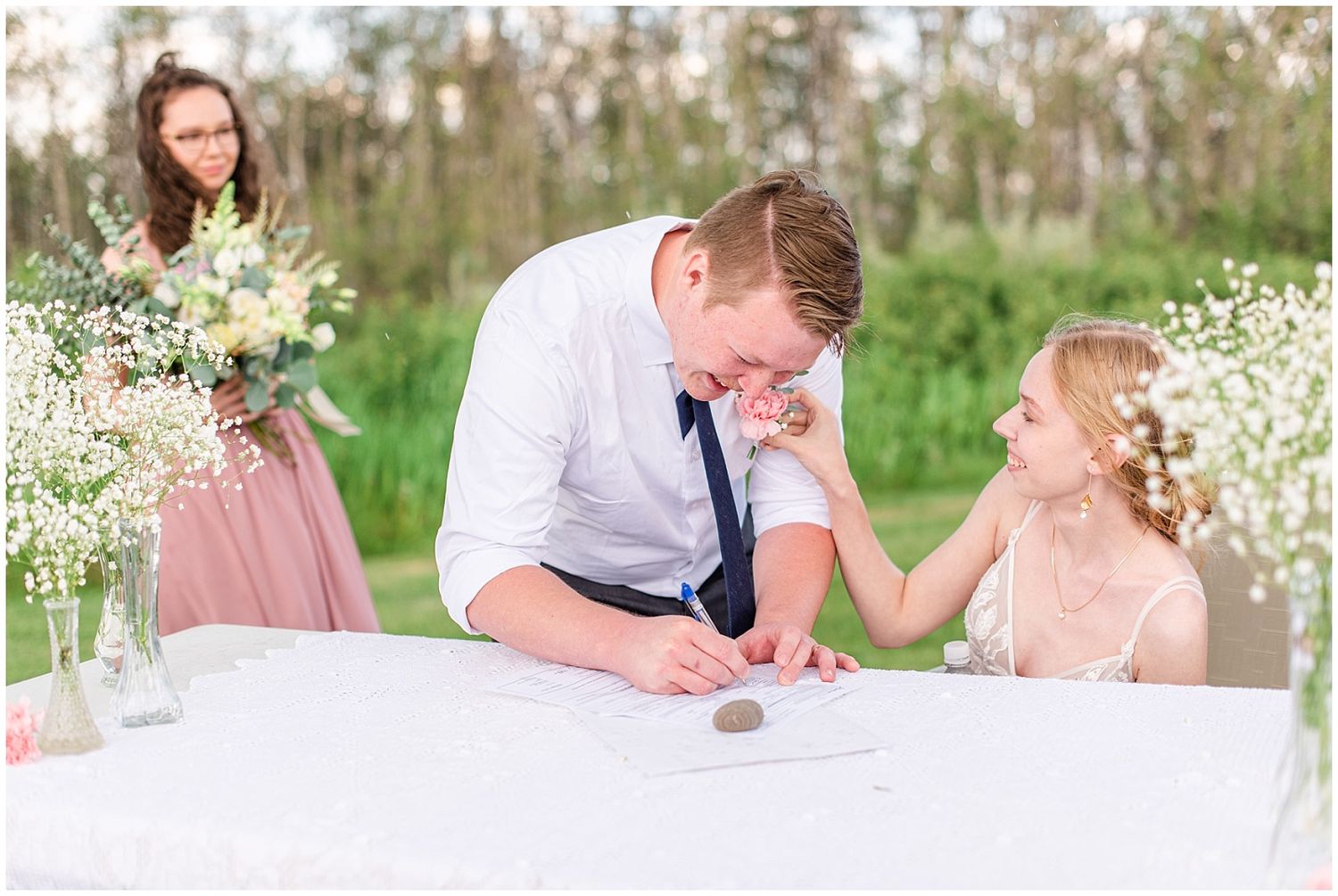 Groom signing wedding documents at table while bride smiles with bridesmaid holding bouquet behind.
