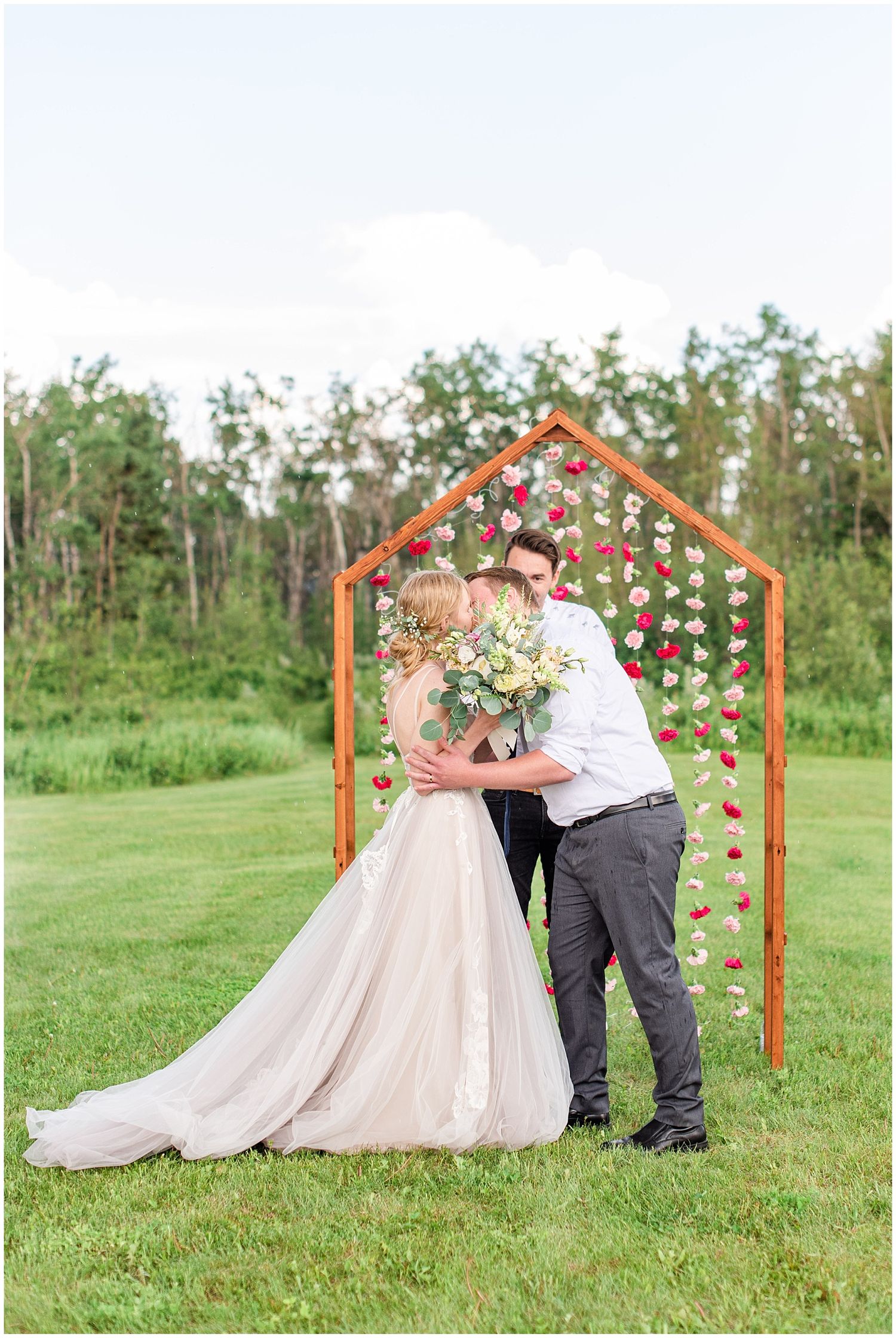Bride and groom share a first kiss under a wooden arch decorated with hanging red and pink flowers on a green lawn.