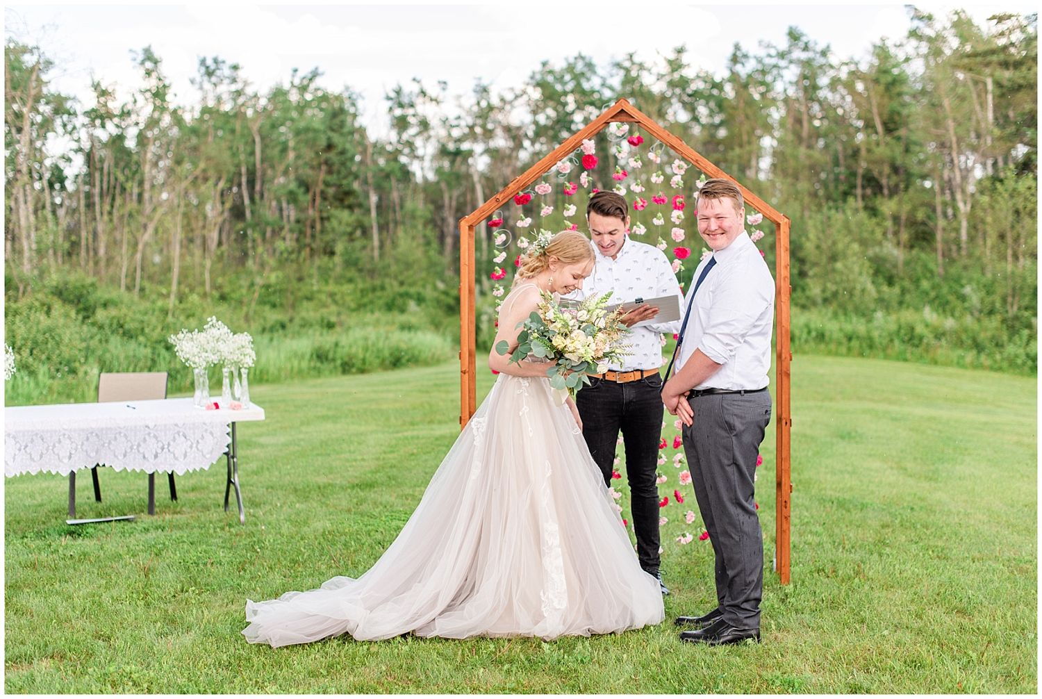 Bride holding bouquet stands with groom and officiant at floral arch after ceremony.
