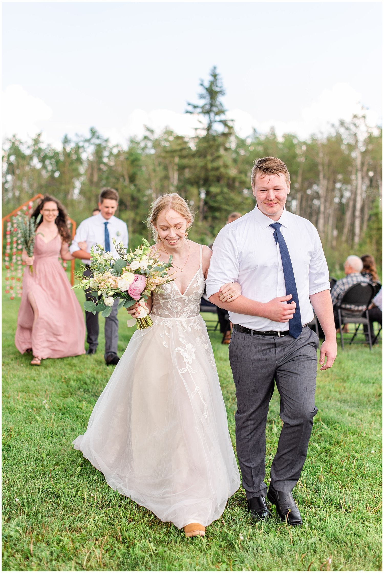 Newlywed couple walking down aisle after outdoor wedding ceremony with wedding party following behind.