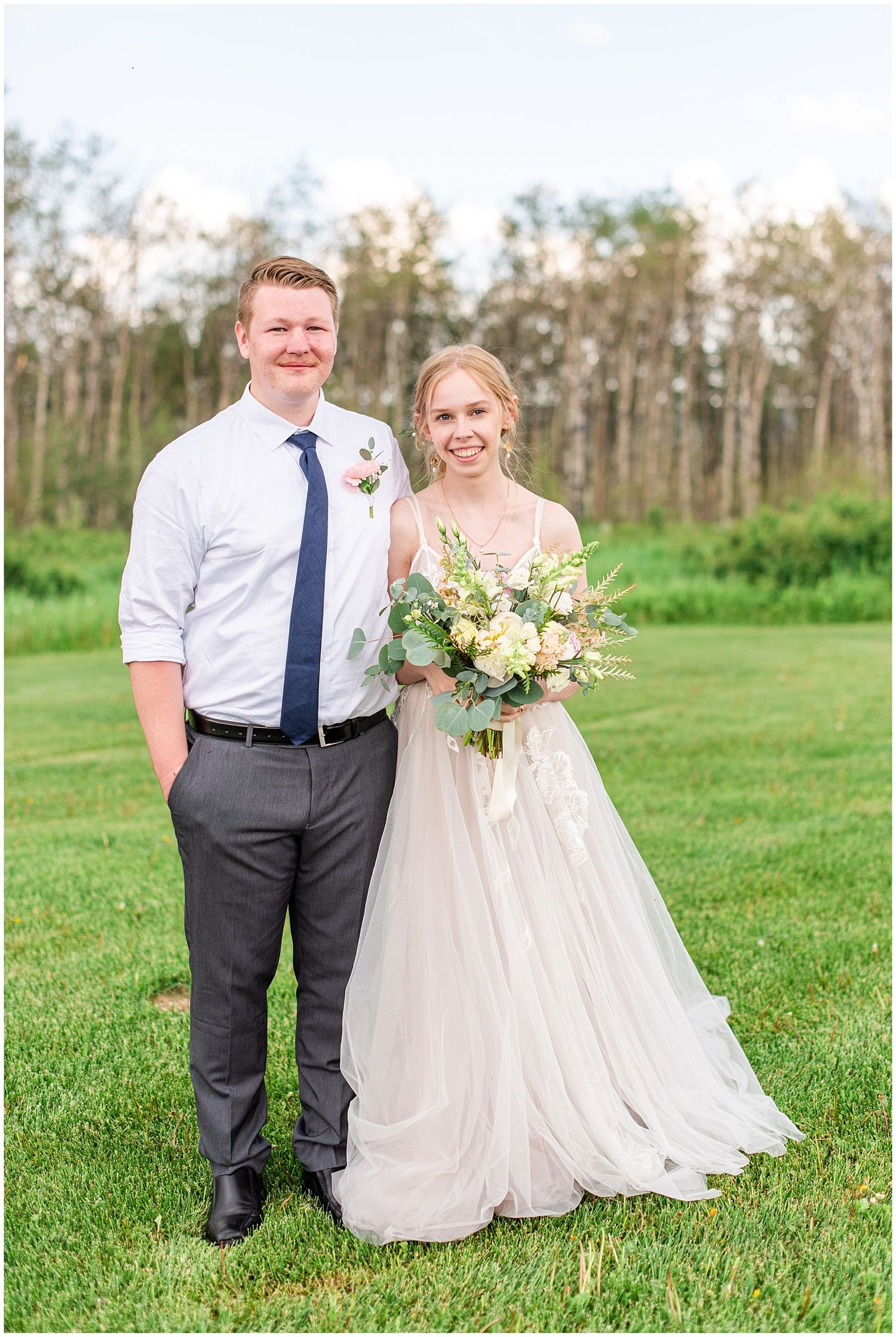Bride in white gown holding floral bouquet stands with groom in gray suit and navy tie on green lawn.