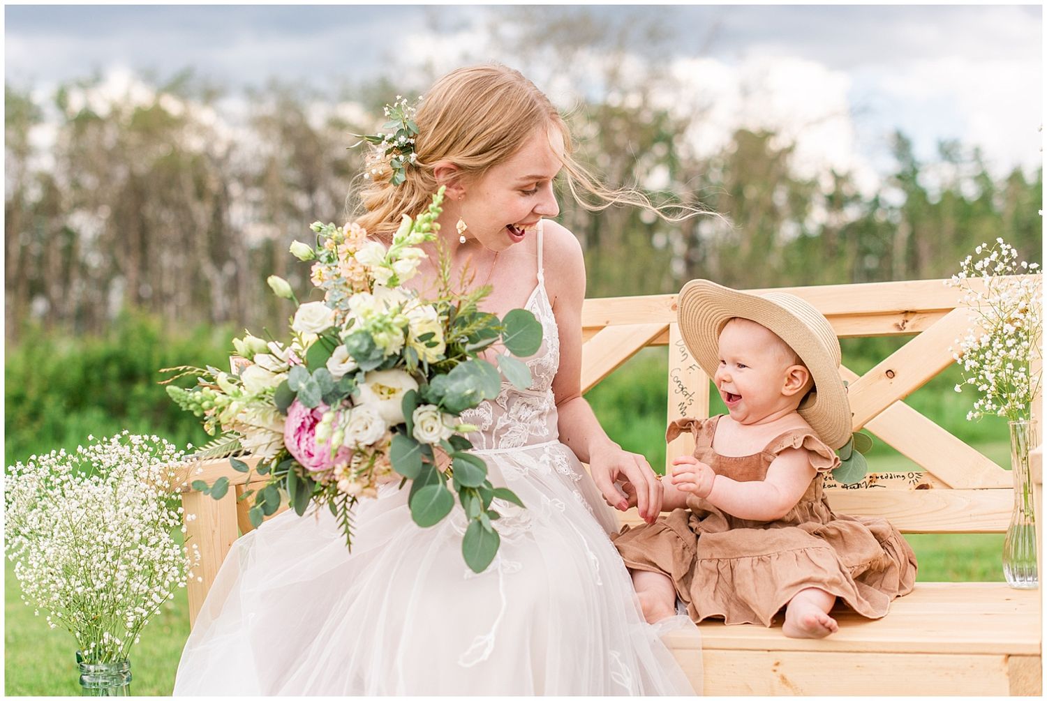 Bride in white gown holding floral bouquet smiles at baby in sun hat seated on wooden bench outdoors.