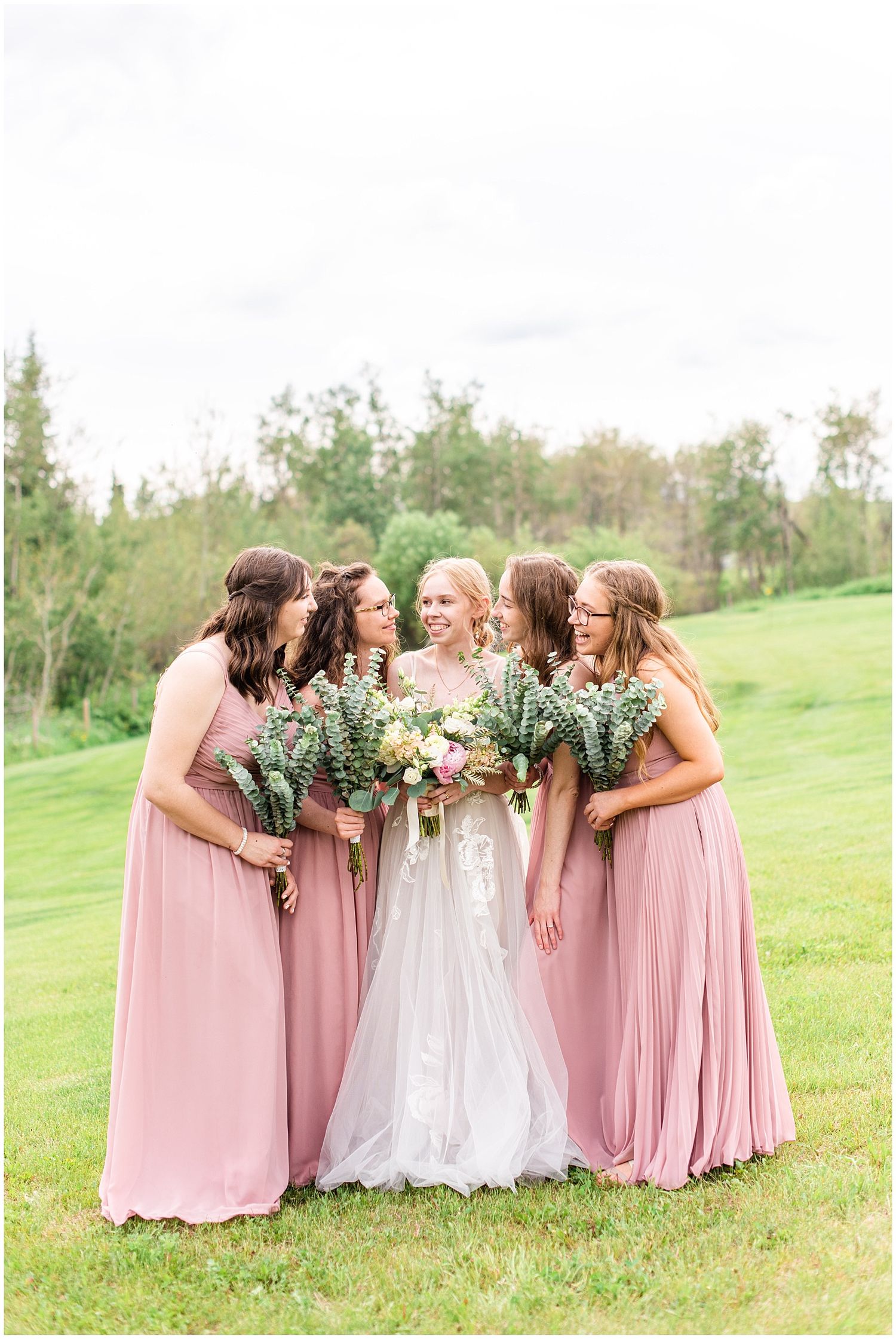 Bride in white lace gown surrounded by four bridesmaids in dusty rose dresses holding greenery bouquets outdoors.
