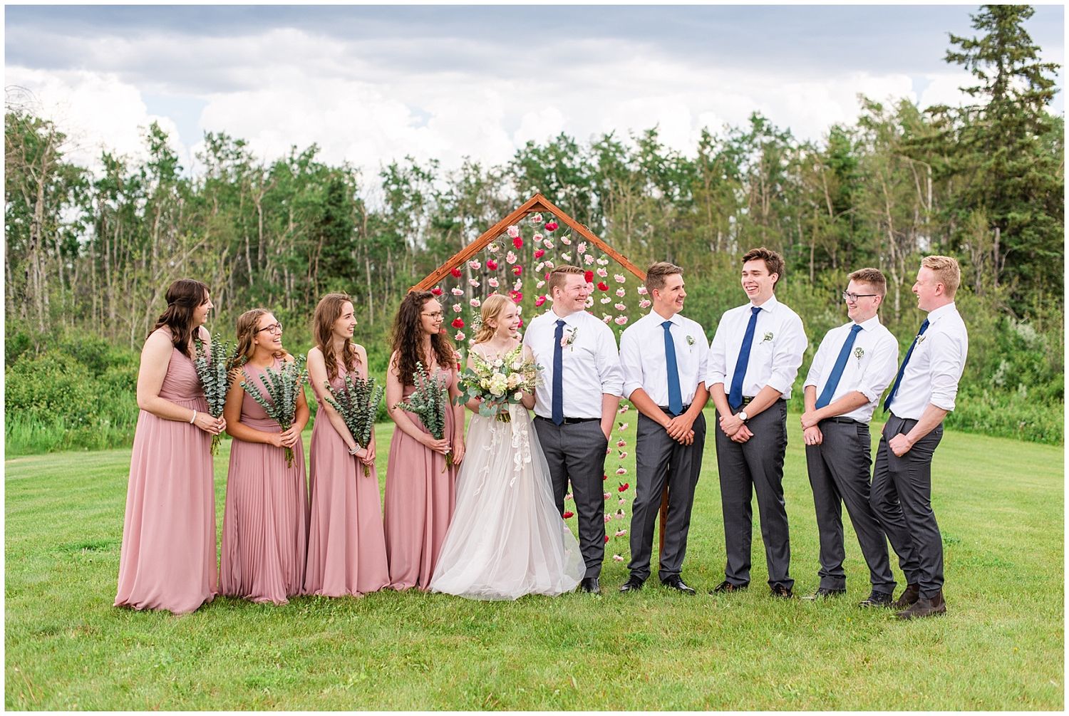 Wedding party posing in front of wooden floral arch with bridesmaids in pink and groomsmen in white.