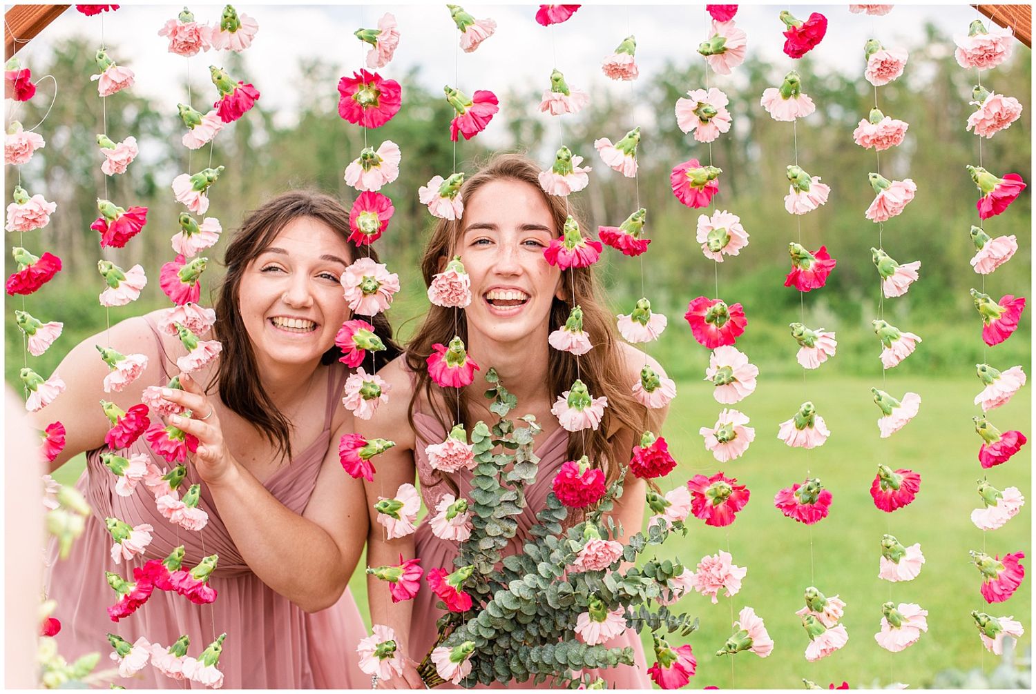 Two bridesmaids laugh joyfully surrounded by hanging pink and red floral curtain backdrop at outdoor wedding celebration.