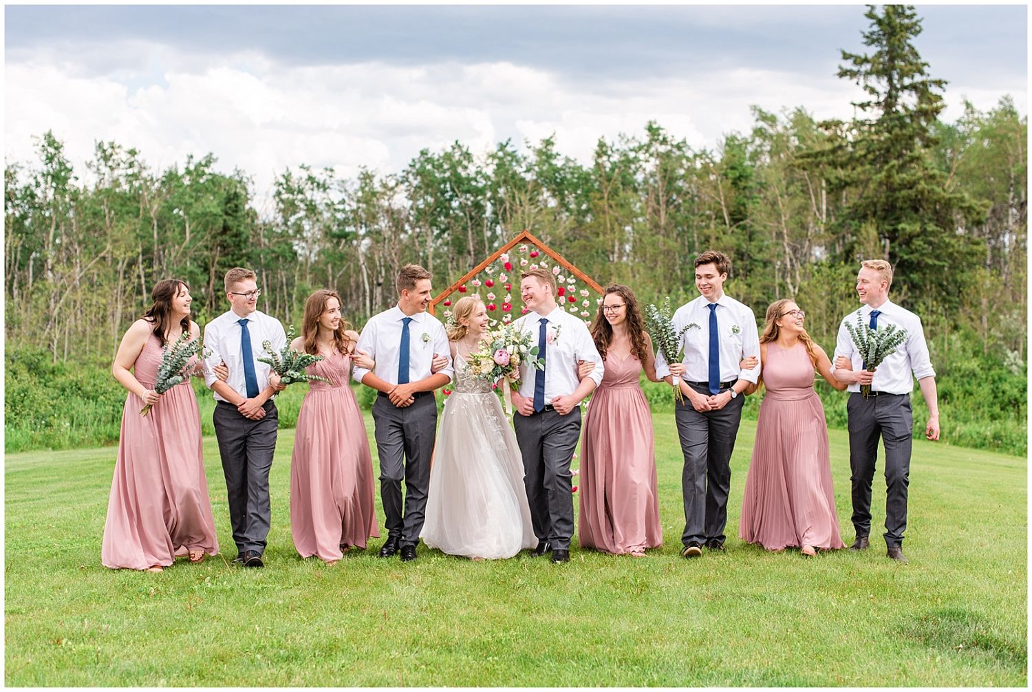 Wedding party in dusty rose dresses and navy ties stands together in front of a floral wooden arch outdoors.