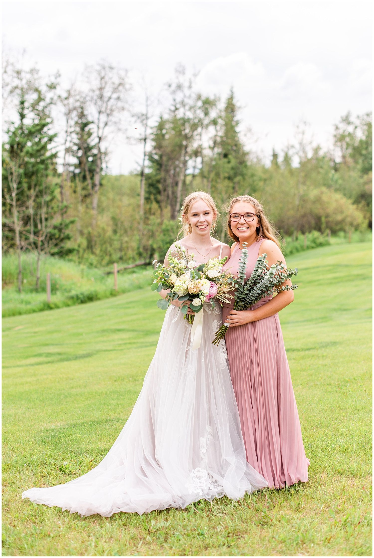 Bride and bridesmaid in dusty rose pleated dress smiling together outdoors holding floral bouquets on green lawn.