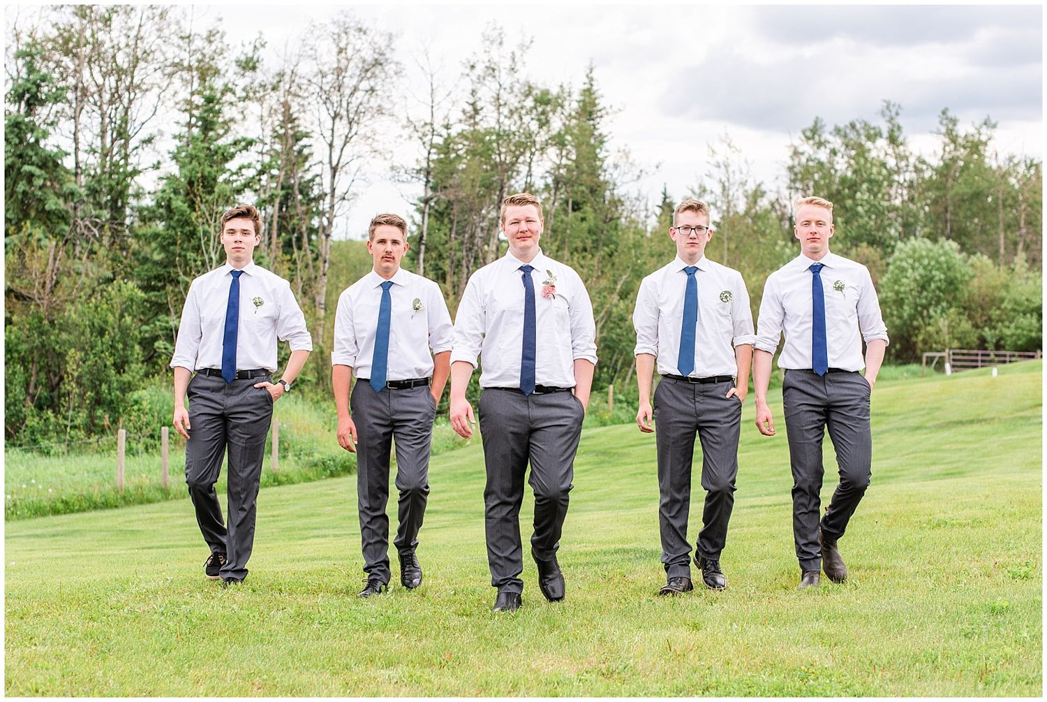 Five groomsmen in white shirts and navy ties walk across green lawn at wedding.