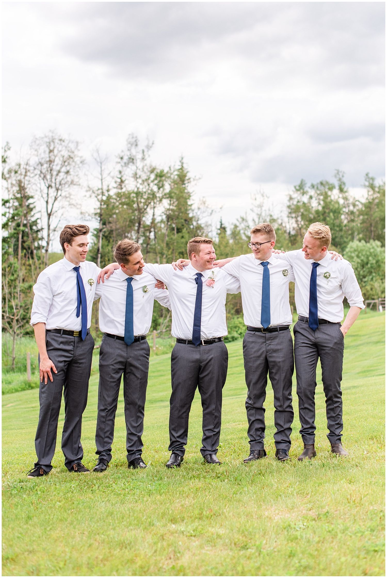 Five groomsmen in white shirts and blue ties laughing together on a green lawn with trees in the background.