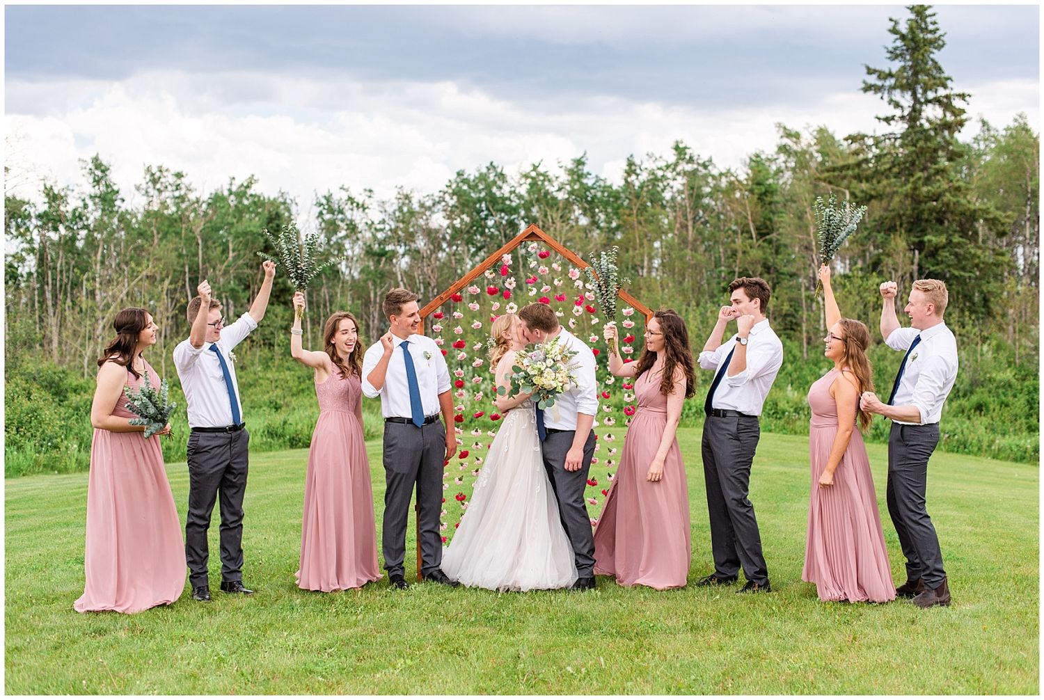 Wedding party celebrating with raised arms as bride and groom kiss before decorative wooden arch.