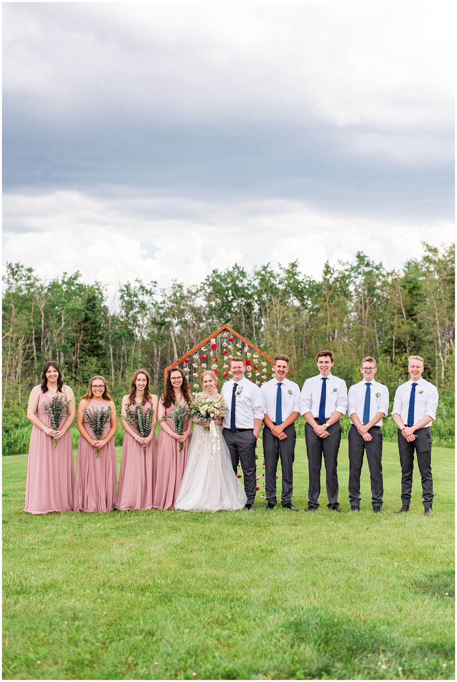 Wedding party poses outdoors on green lawn, bridesmaids in pink gowns and groomsmen in gray suits with navy ties.
